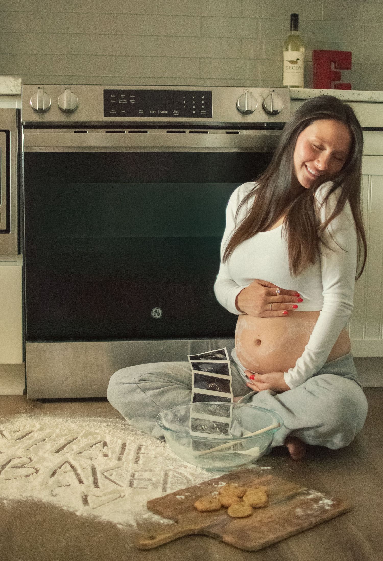 Smiling pregnant woman sitting on kitchen floor with baby name written in flour near ultrasound photos.