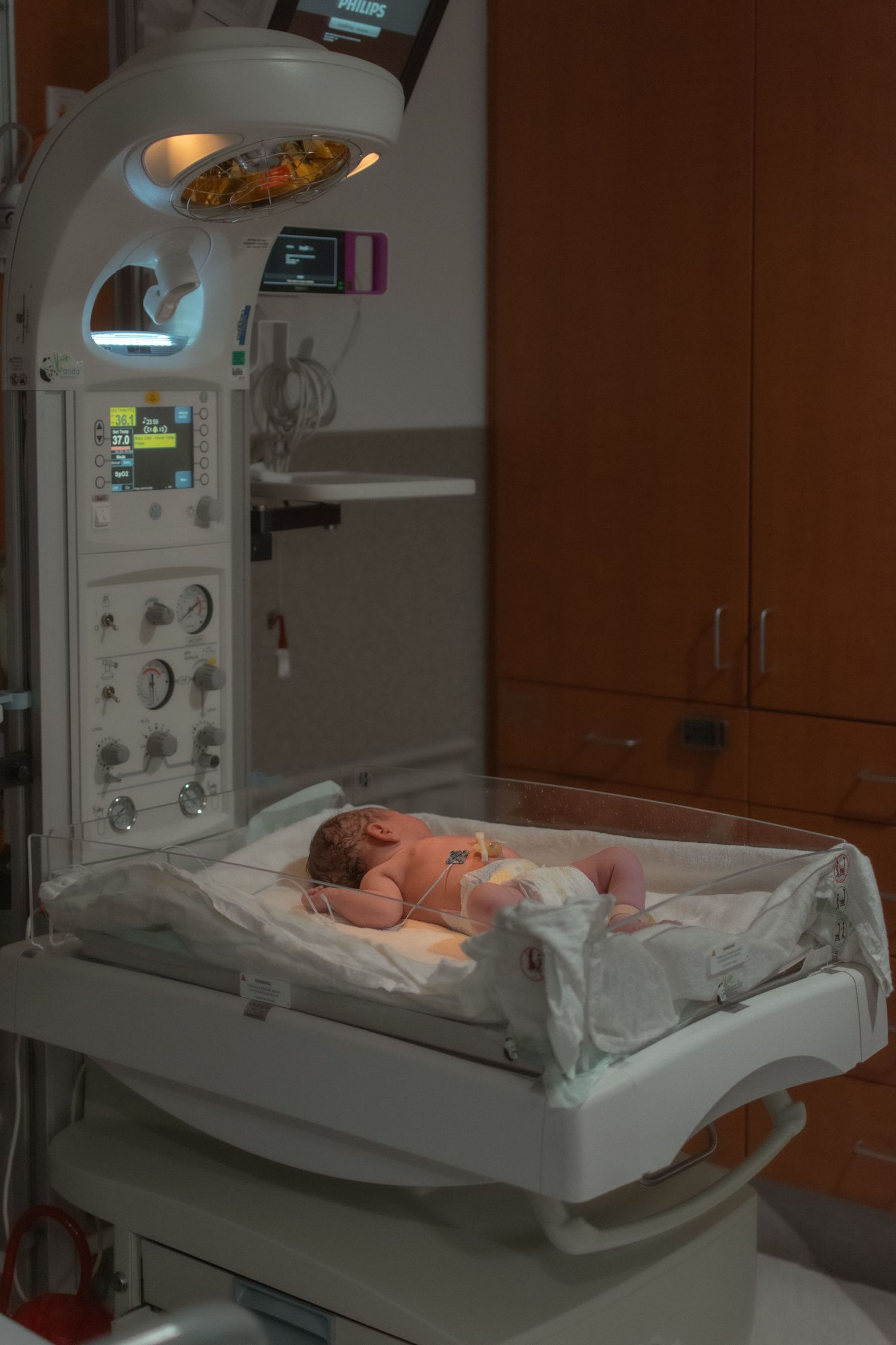 Newborn baby lying in a hospital incubator under warming lights in a neonatal care unit.