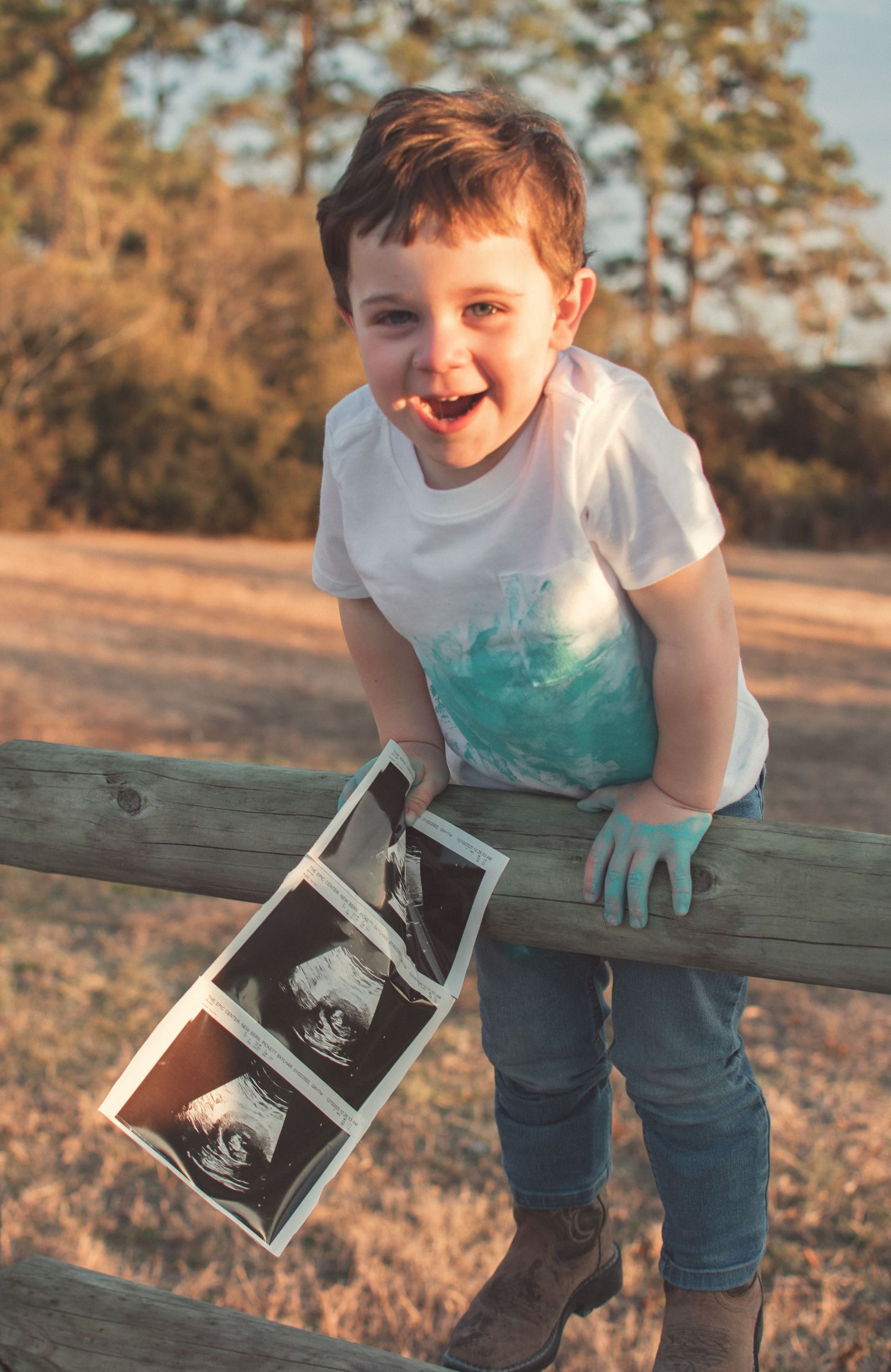 Excited young boy sitting on fence holding ultrasound photos announcing new baby sibling arrival.