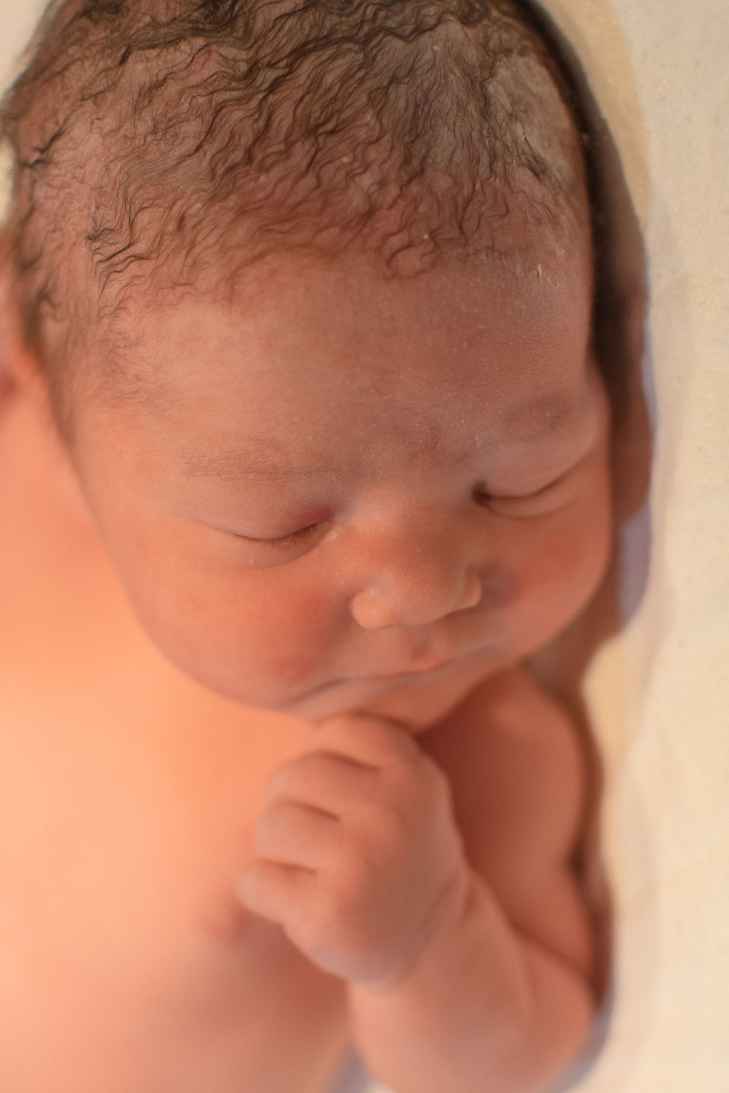 Peaceful newborn baby sleeping with hand near face, featuring curly dark hair and soft skin in warm light.