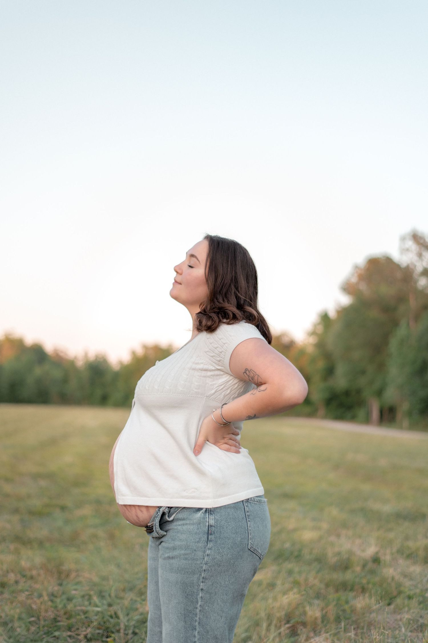 Pregnant woman in white top and jeans stands in a sunlit field, smiling with eyes closed, hand on back.