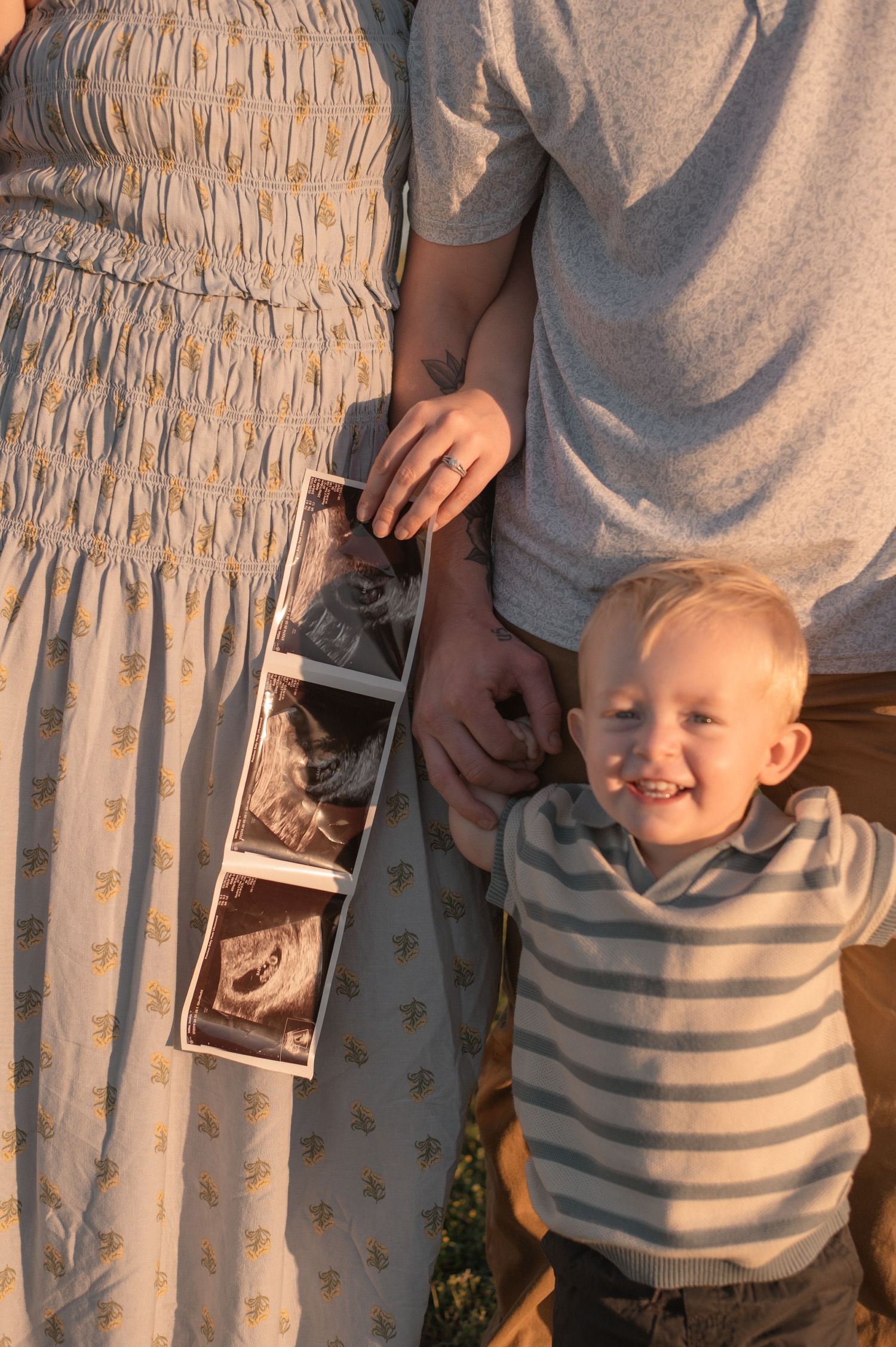 A young couple holds ultrasound photos while their smiling toddler boy stands beside them during a golden hour maternity photo session.