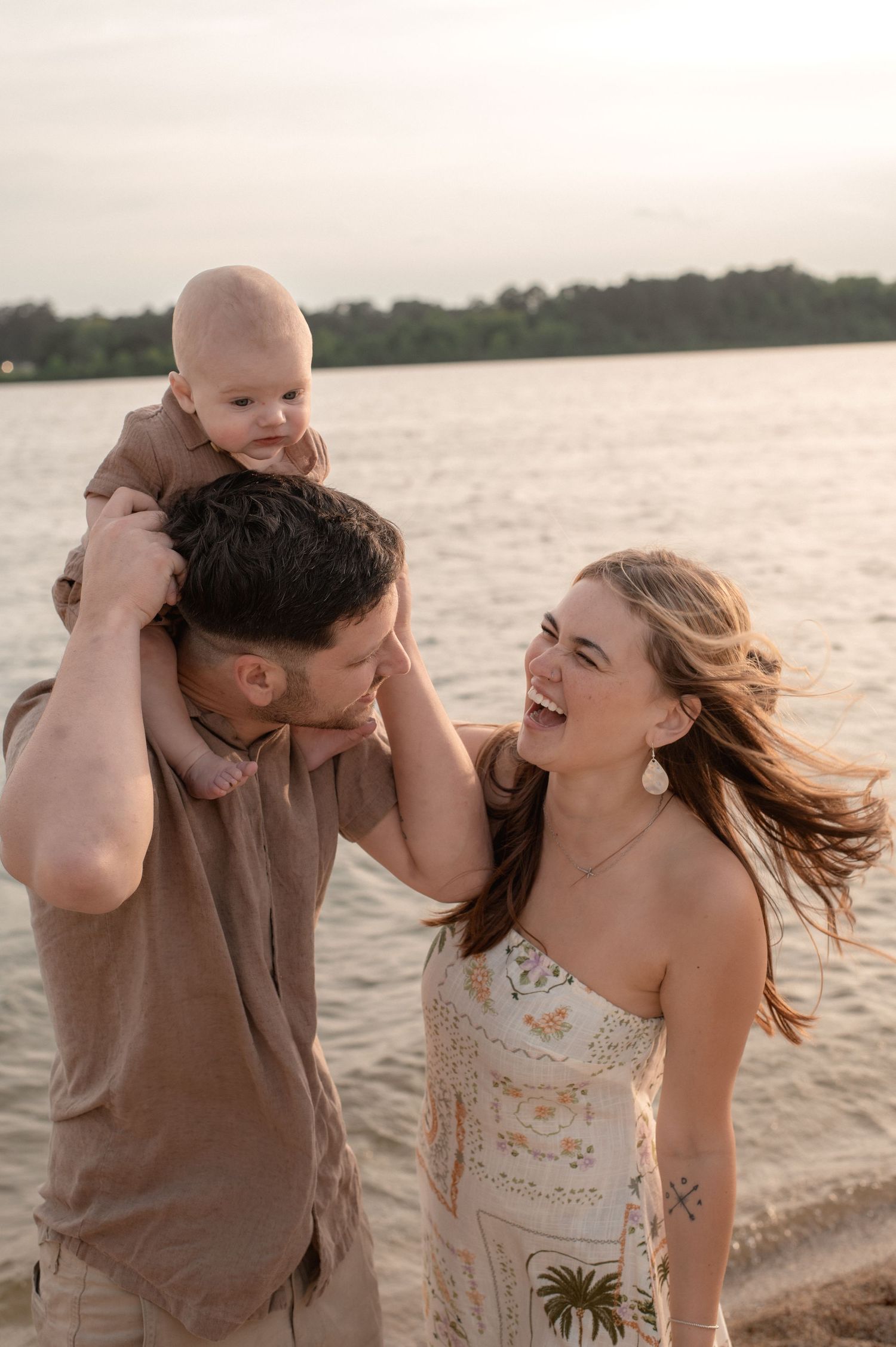 Happy young family laughing on beach at sunset, father holding baby on shoulders, mother in floral dress smiling.