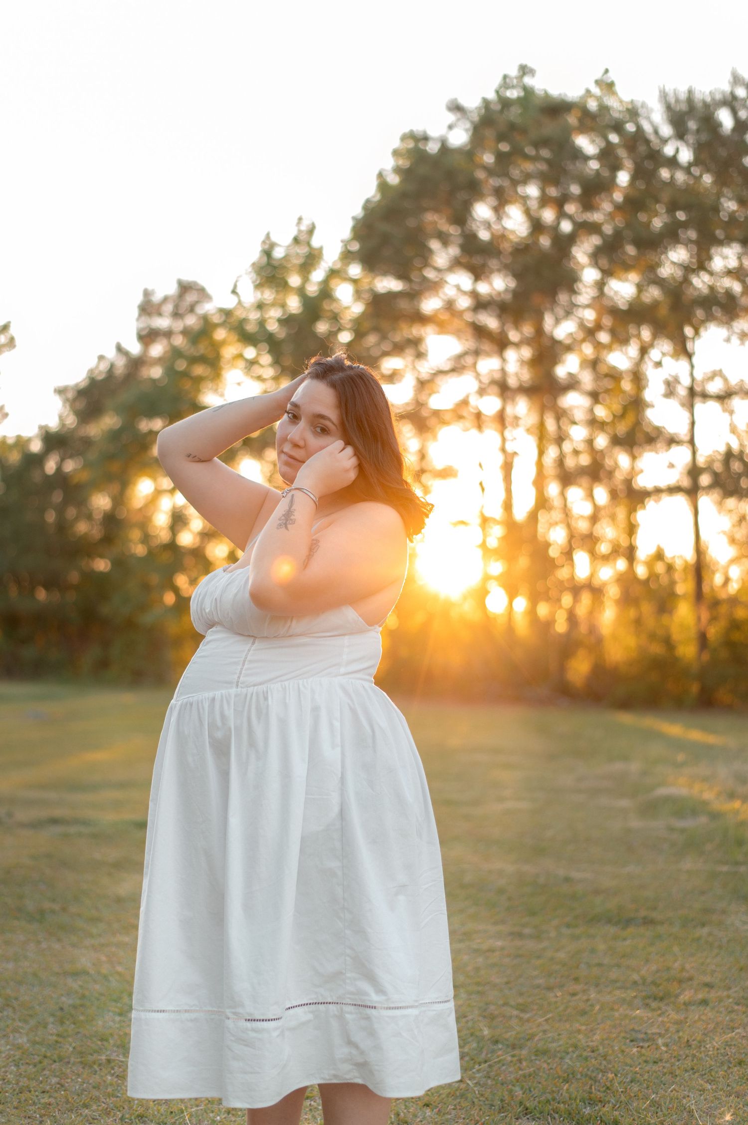 Woman in white sundress posing at golden hour sunset in a green field with tall trees in background.