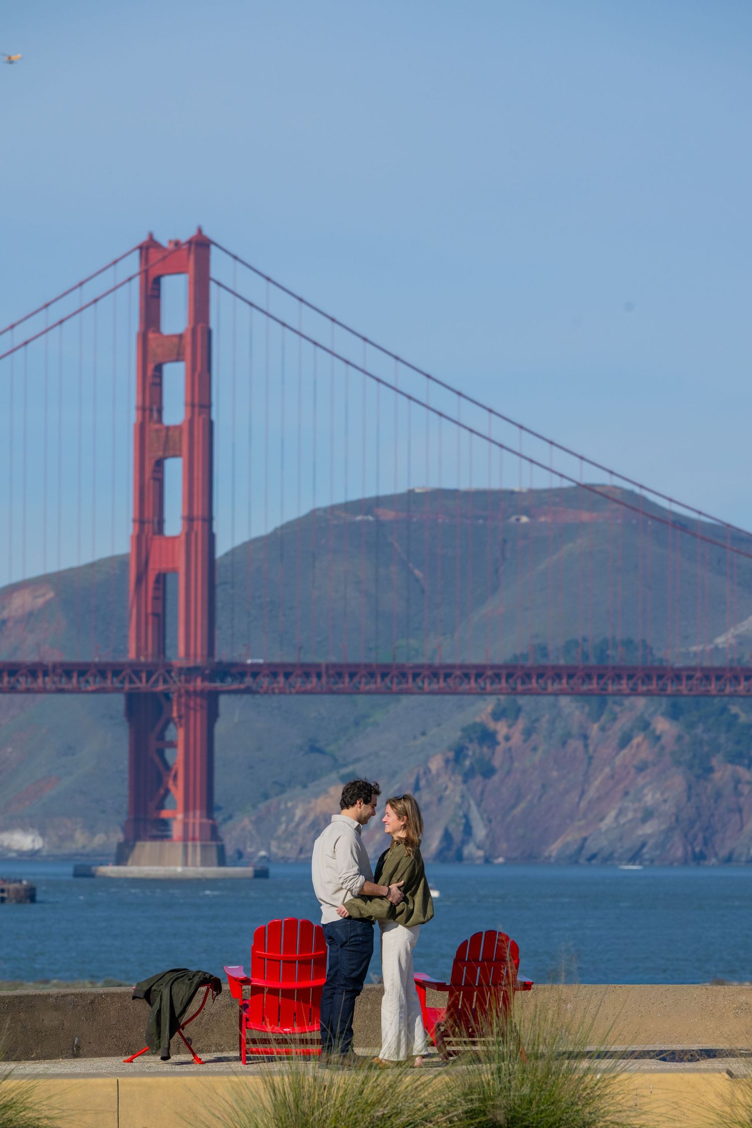 Couple embracing in front of the iconic Golden Gate Bridge in San Francisco, with red Adirondack chairs nearby.
