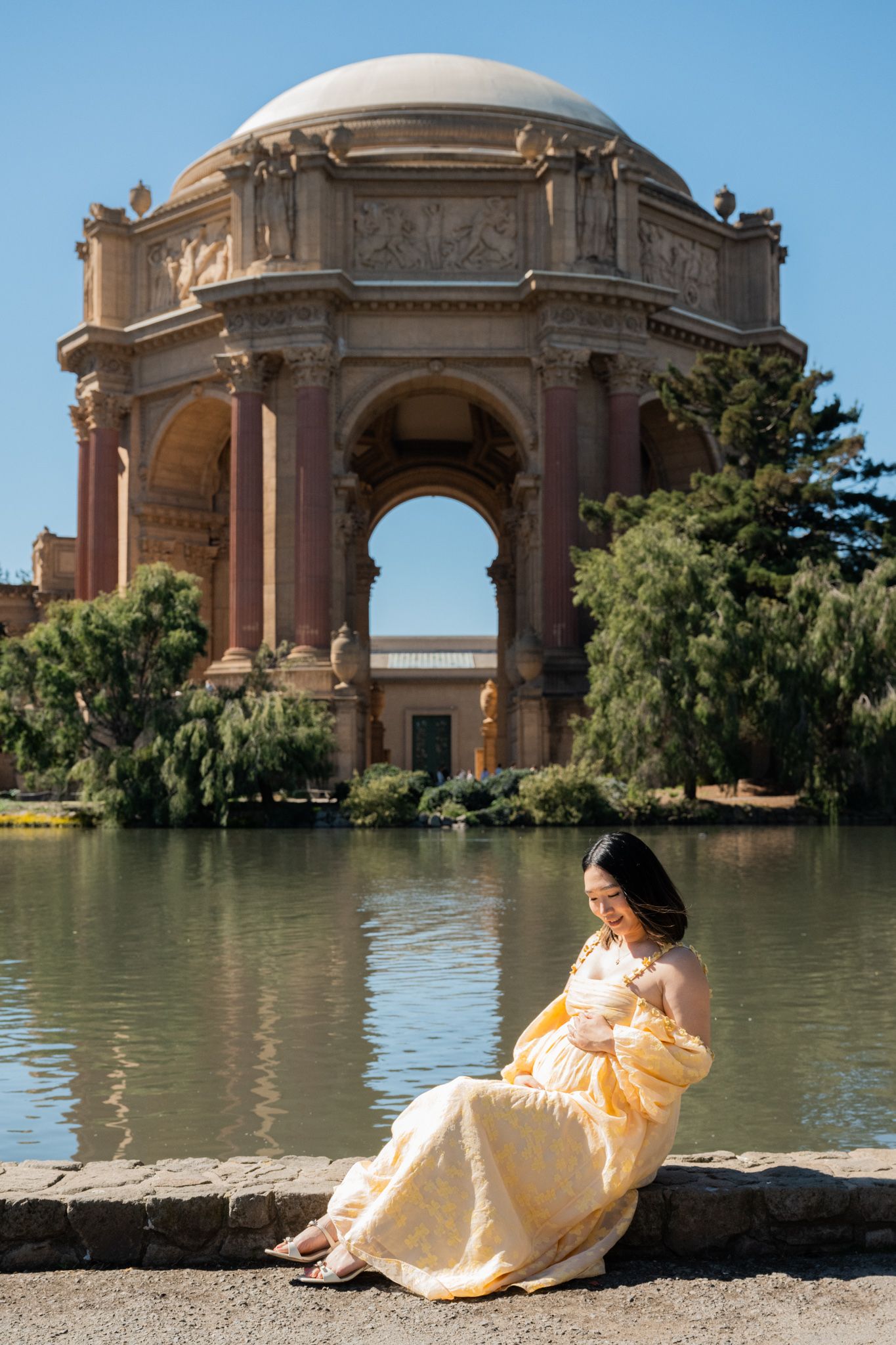 Pregnant woman in yellow gown sits by lagoon at Palace of Fine Arts, San Francisco, on a sunny day.