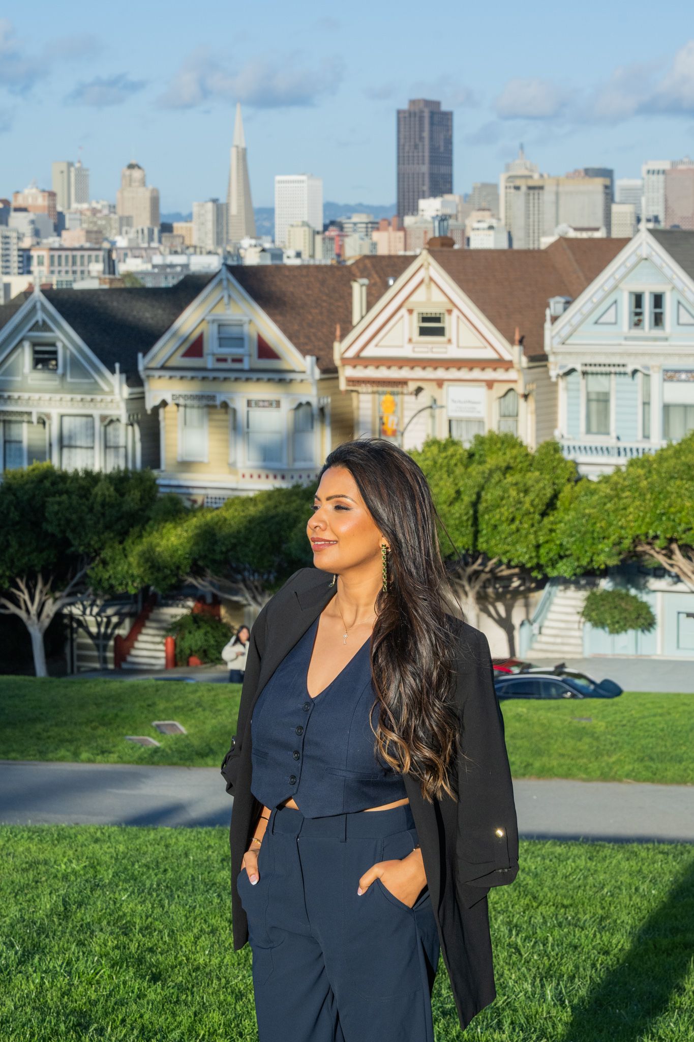 Woman in navy blue outfit stands at Alamo Square with the Painted Ladies and SF skyline behind her.