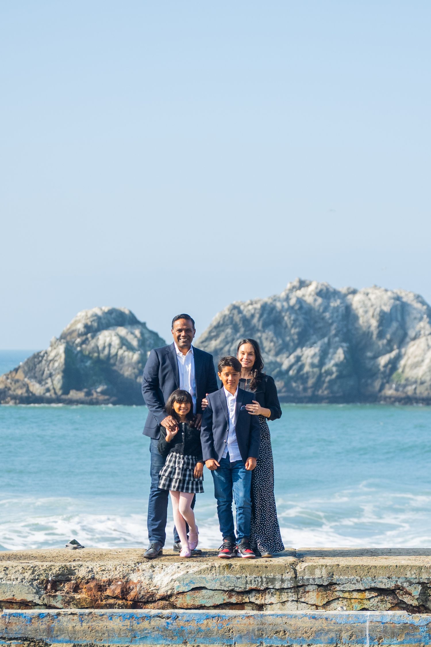 A family of four poses on a seaside wall with rocky cliffs and ocean waves in the background.