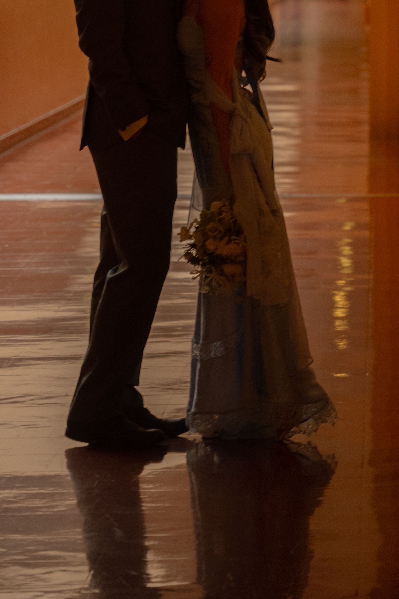 Bride in flowing gown holds bouquet while standing close to groom on reflective floor.