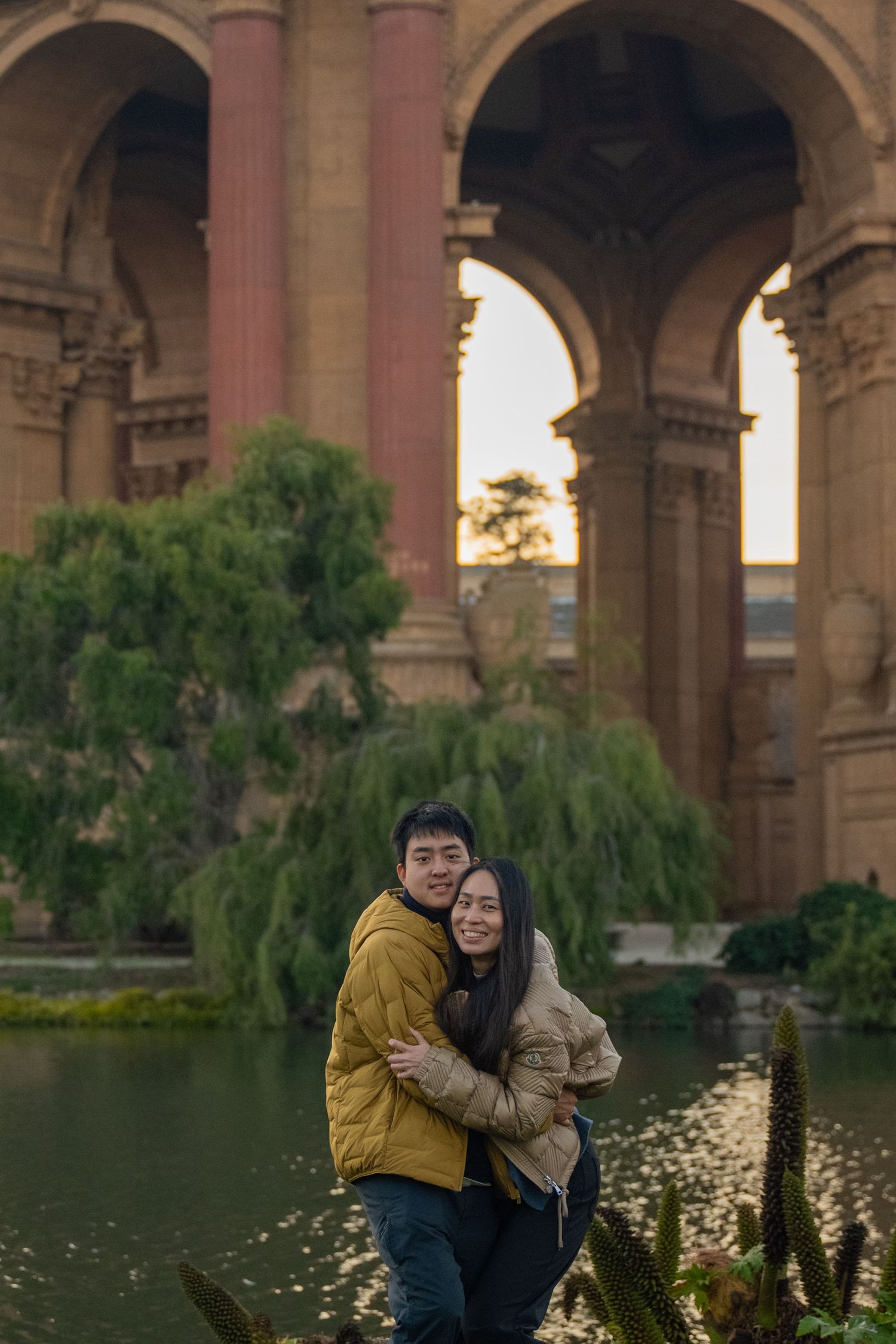 A couple embraces and smiles in front of the Palace of Fine Arts arches and pond at dusk.