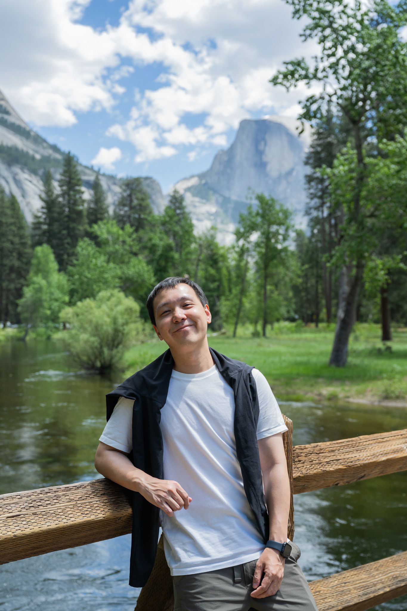 Smiling Asian man leans on wooden fence with Half Dome and Yosemite Valley in the background.
