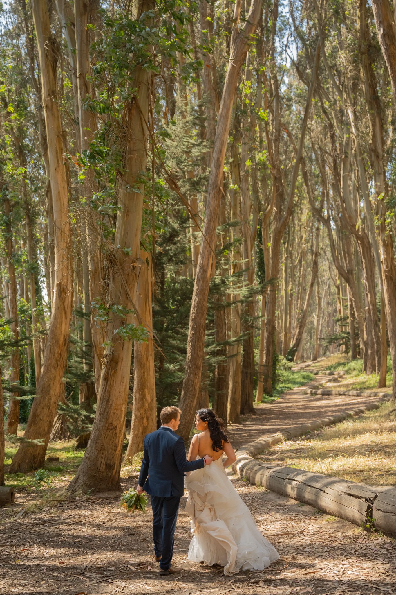 Bride and groom walk hand in hand through a sunlit eucalyptus forest path at their wedding.