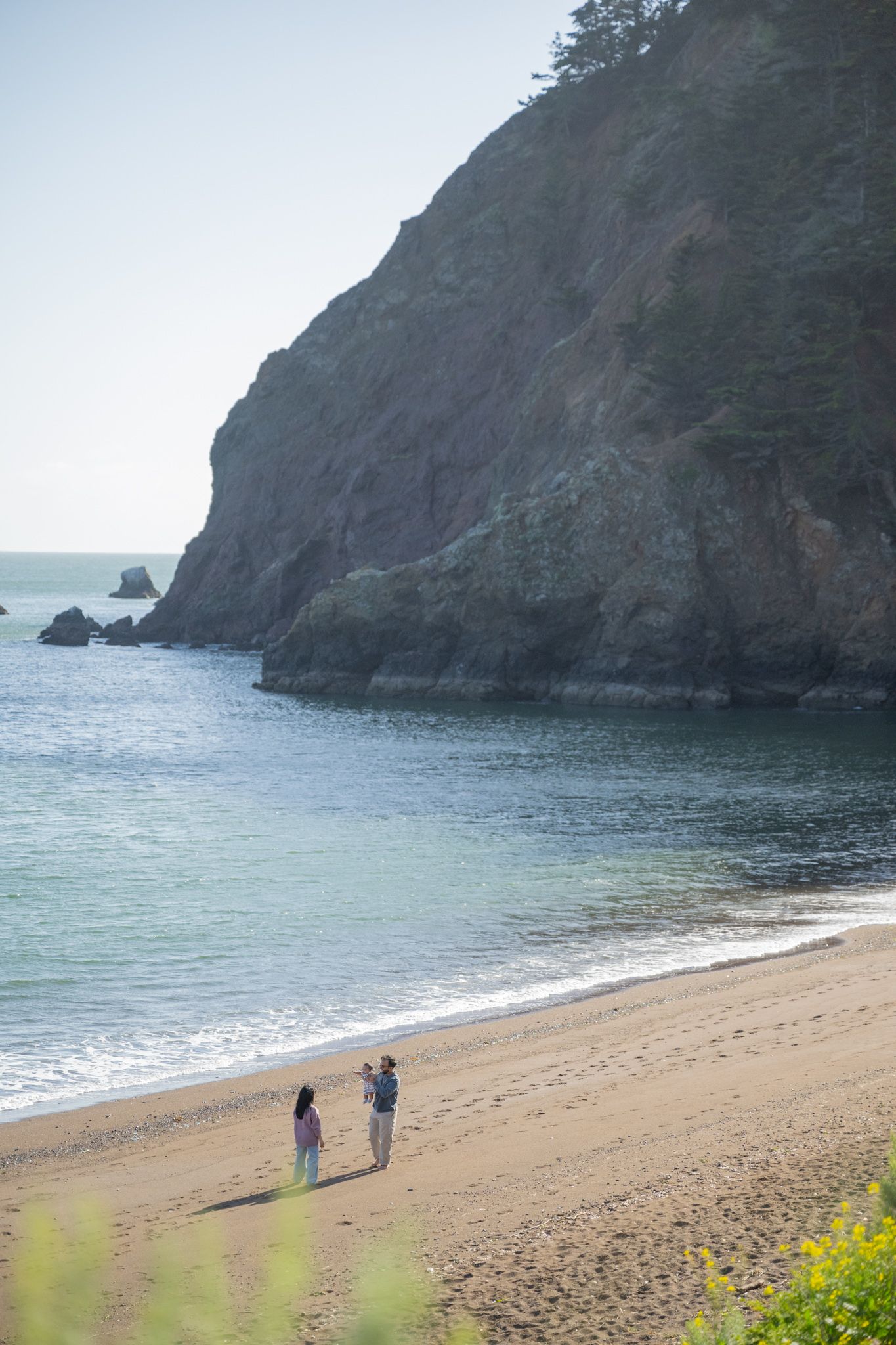 Couple walks hand in hand on a serene sandy beach beside dramatic rocky cliffs and calm blue ocean waters.