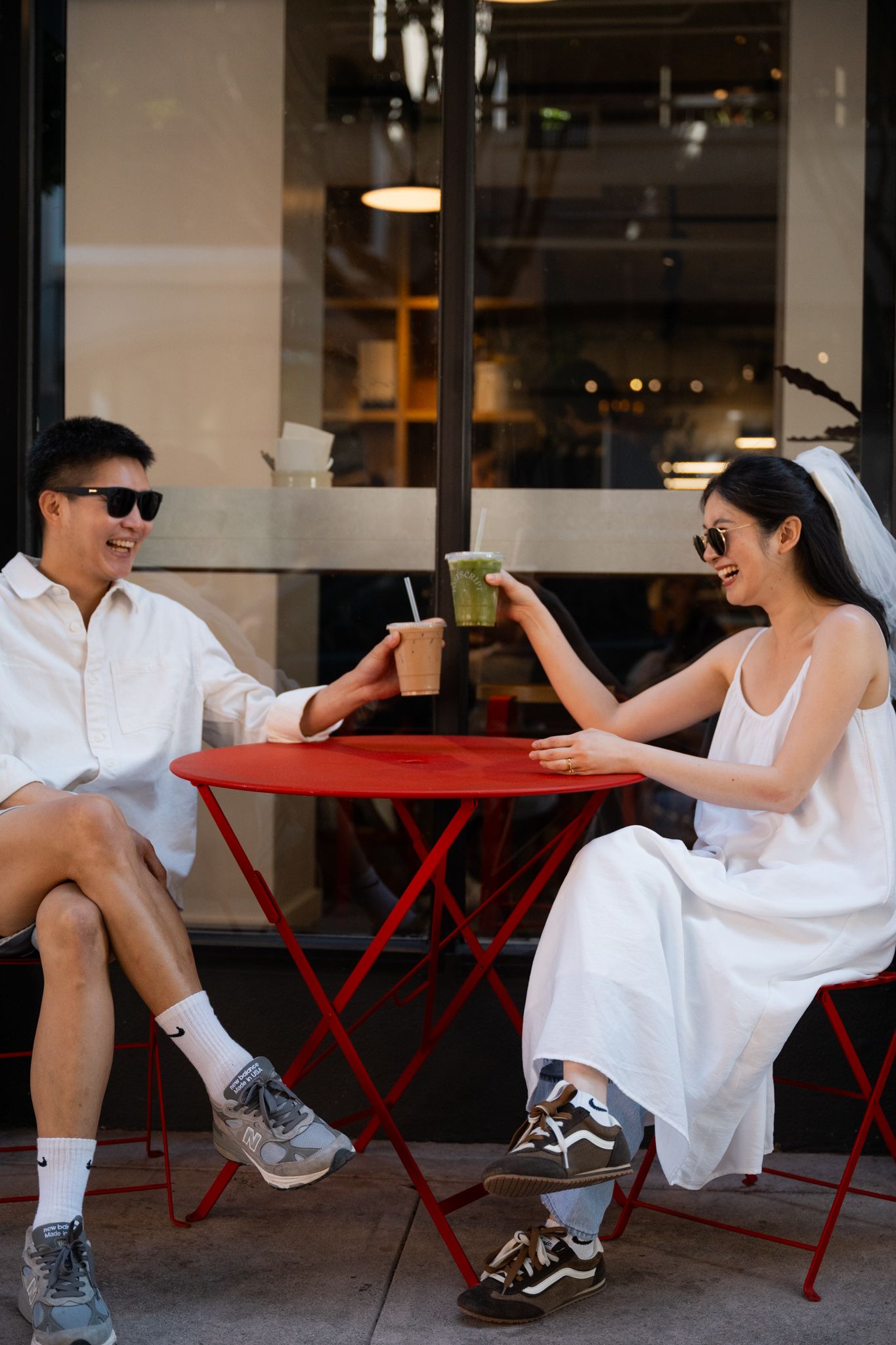 A couple in white outfits toasting drinks at a red outdoor cafe table, both wearing sunglasses and sneakers.