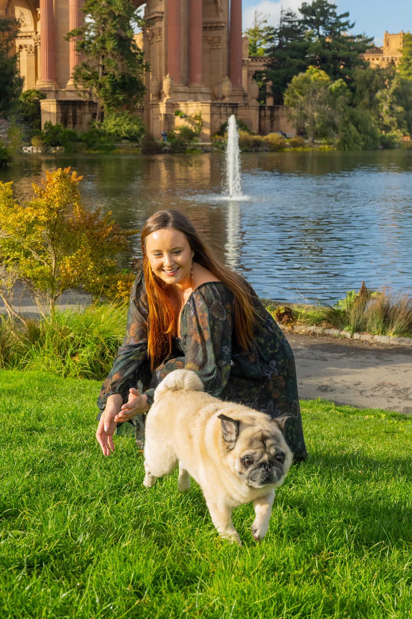 A young woman with long red hair smiles while petting a pug on the grass at the Palace of Fine Arts.