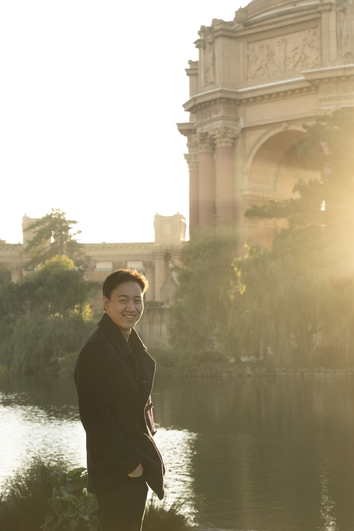 A smiling man in a black jacket stands by a reflective pond near a classical building at sunset.