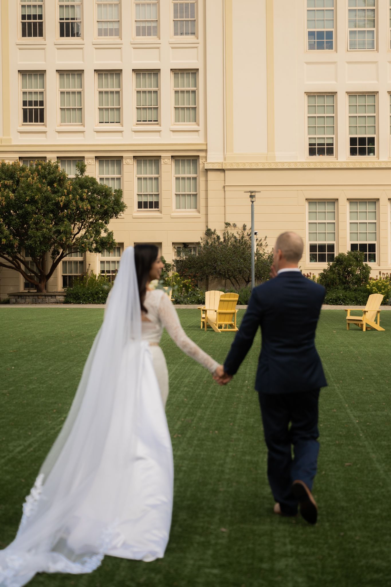 Bride and groom holding hands walking across green lawn toward elegant yellow building with Adirondack chairs.