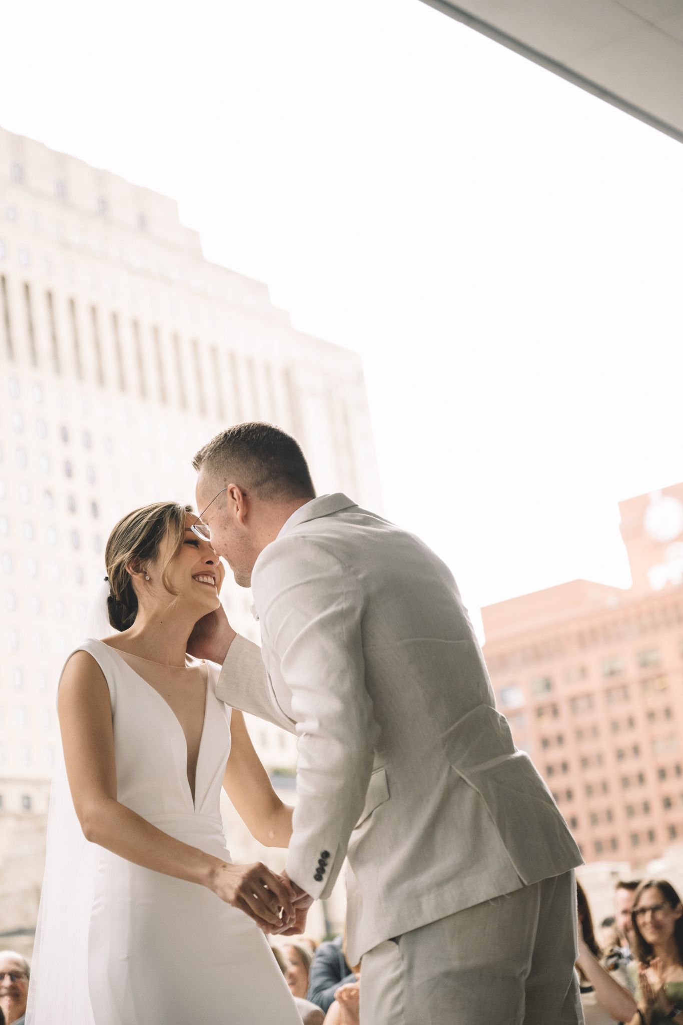 Bride and groom share first kiss during outdoor rooftop wedding ceremony with city skyline backdrop.