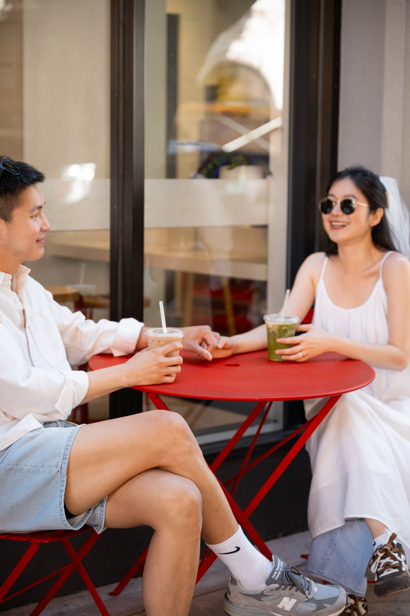 A couple enjoys iced drinks at a red outdoor cafe table, smiling and relaxed on a sunny day.