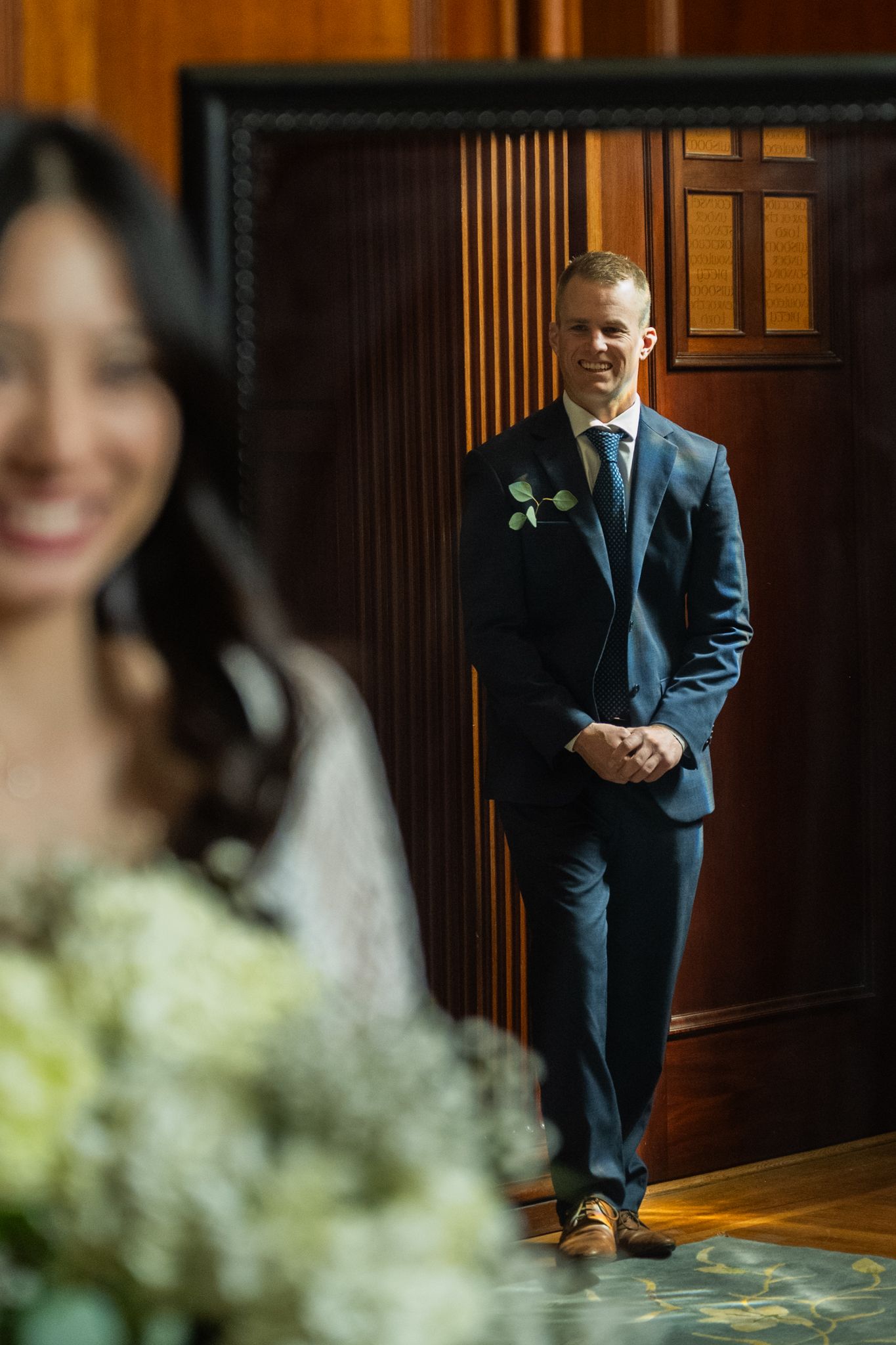 Groom in navy suit with boutonniere stands against wood-paneled wall watching blurred bride in foreground.
