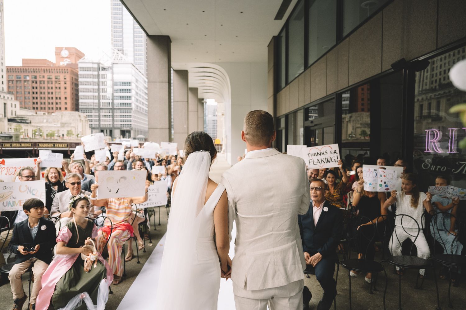 Bride and groom walk toward cheering crowd of guests holding signs outside urban wedding venue.