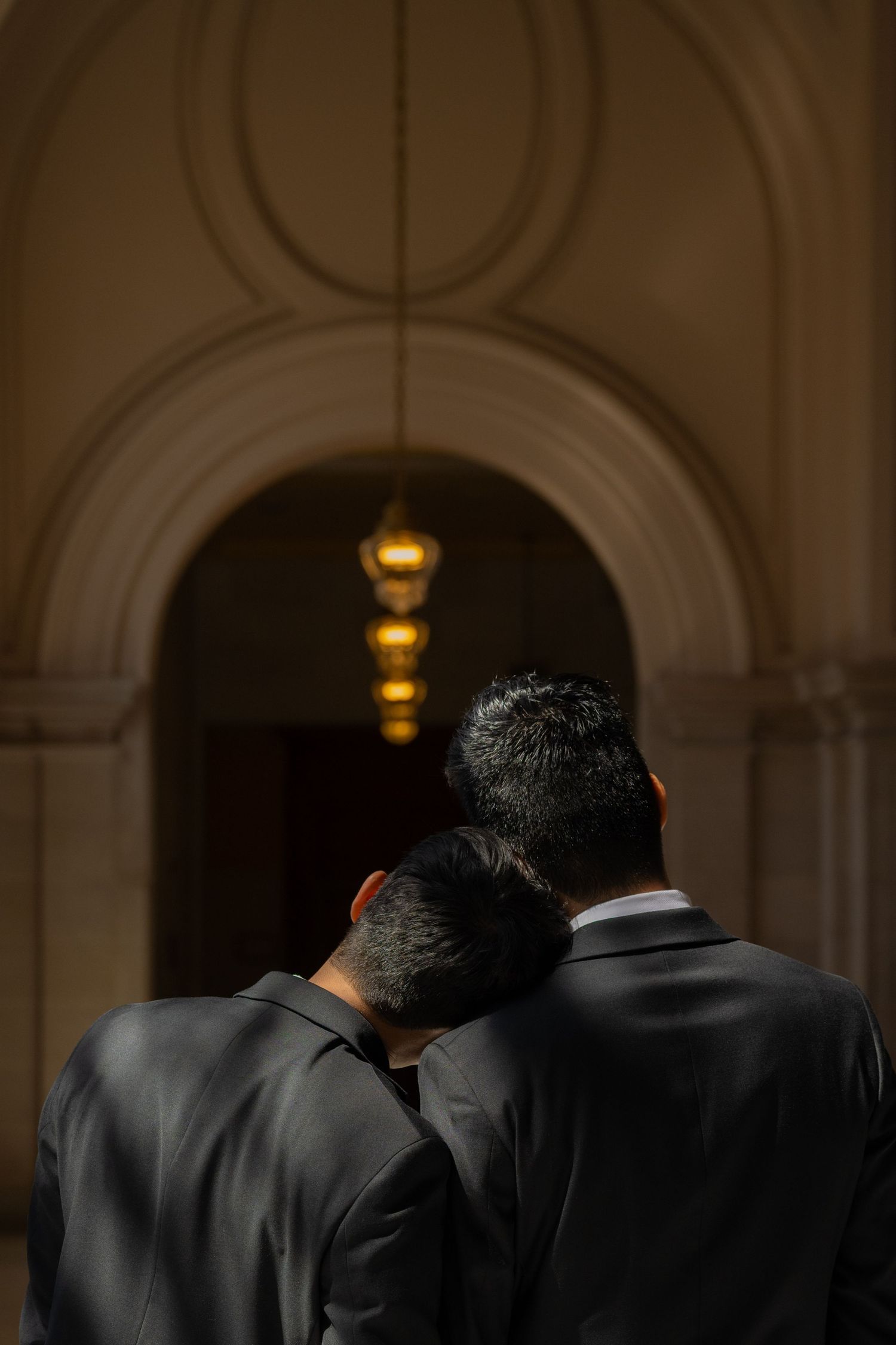 Two wedding guests in black suits lean heads together beneath a glowing chandelier in an arched hallway.