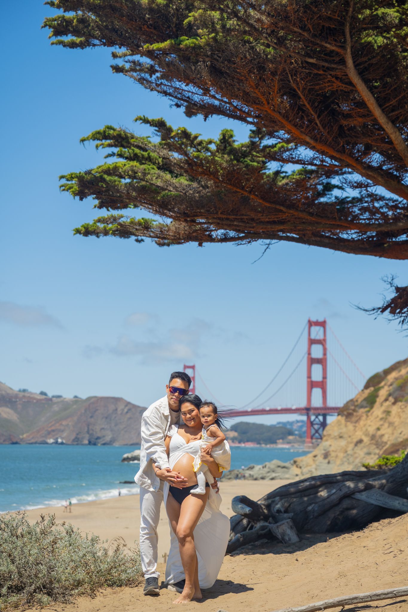 A family of three poses on a sunny San Francisco beach with the iconic Golden Gate Bridge in the background.