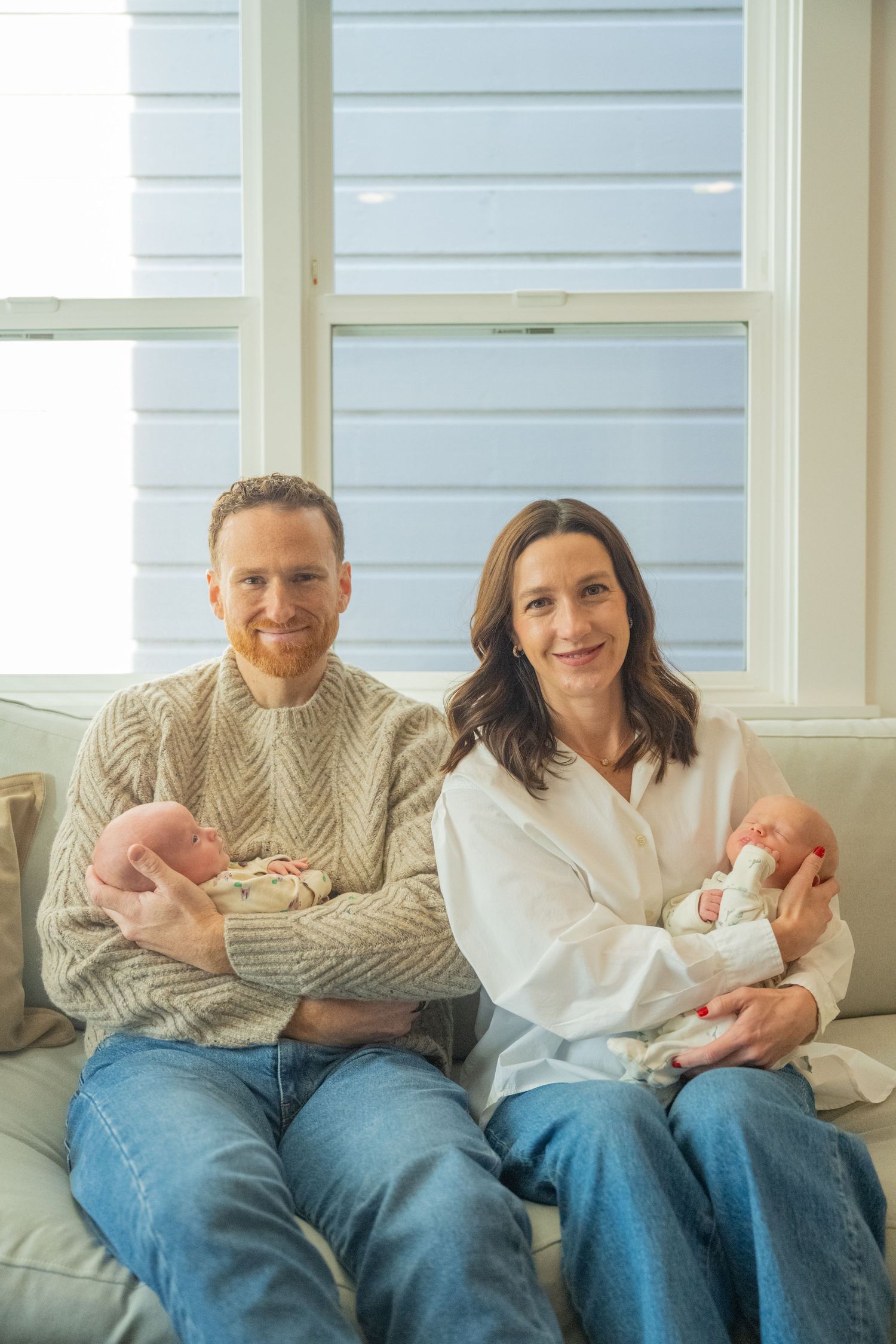 Happy couple sitting on a sofa holding newborn twins, smiling at the camera in a bright home setting.