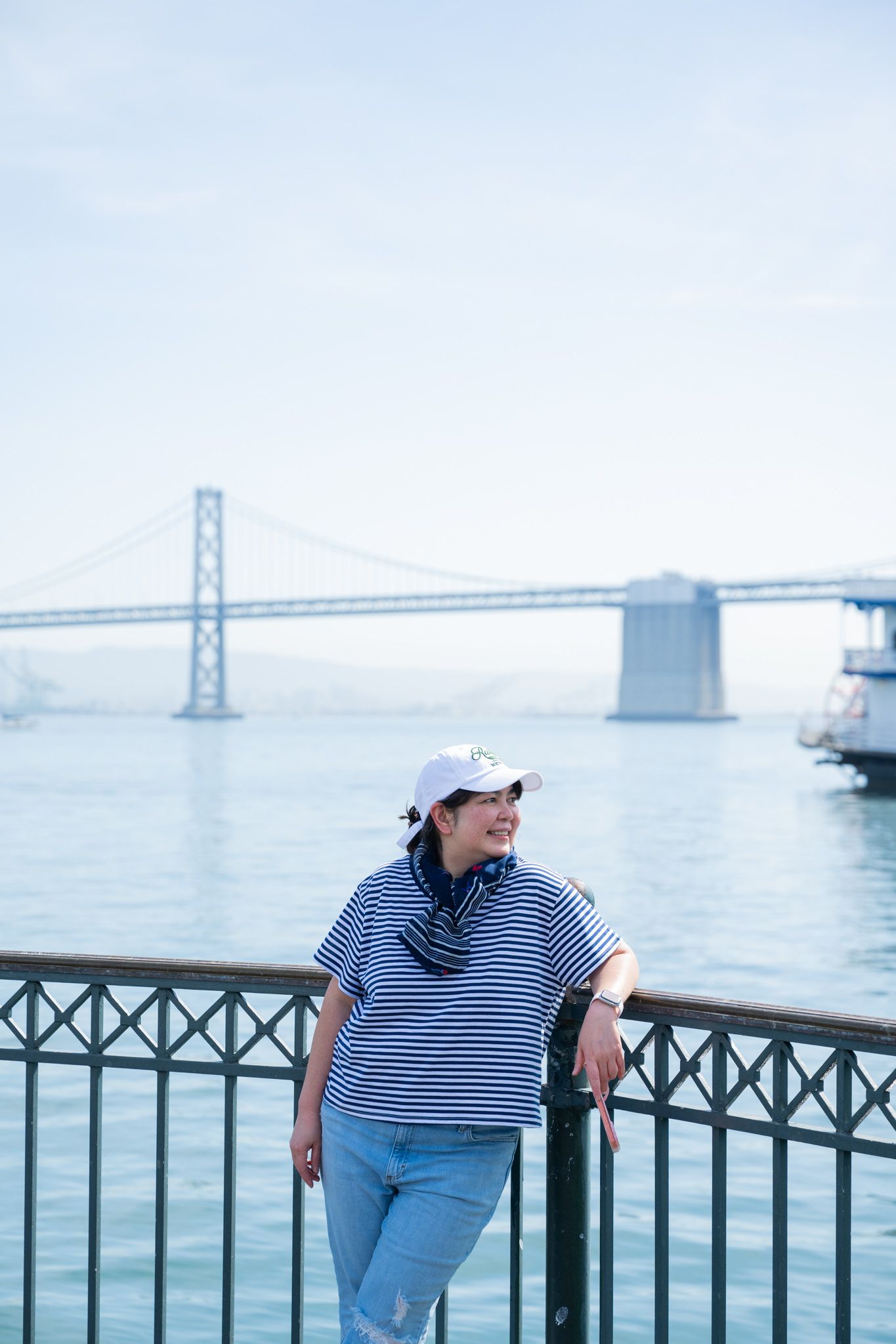 Woman in striped shirt and white cap leans on railing with Bay Bridge visible across the water.