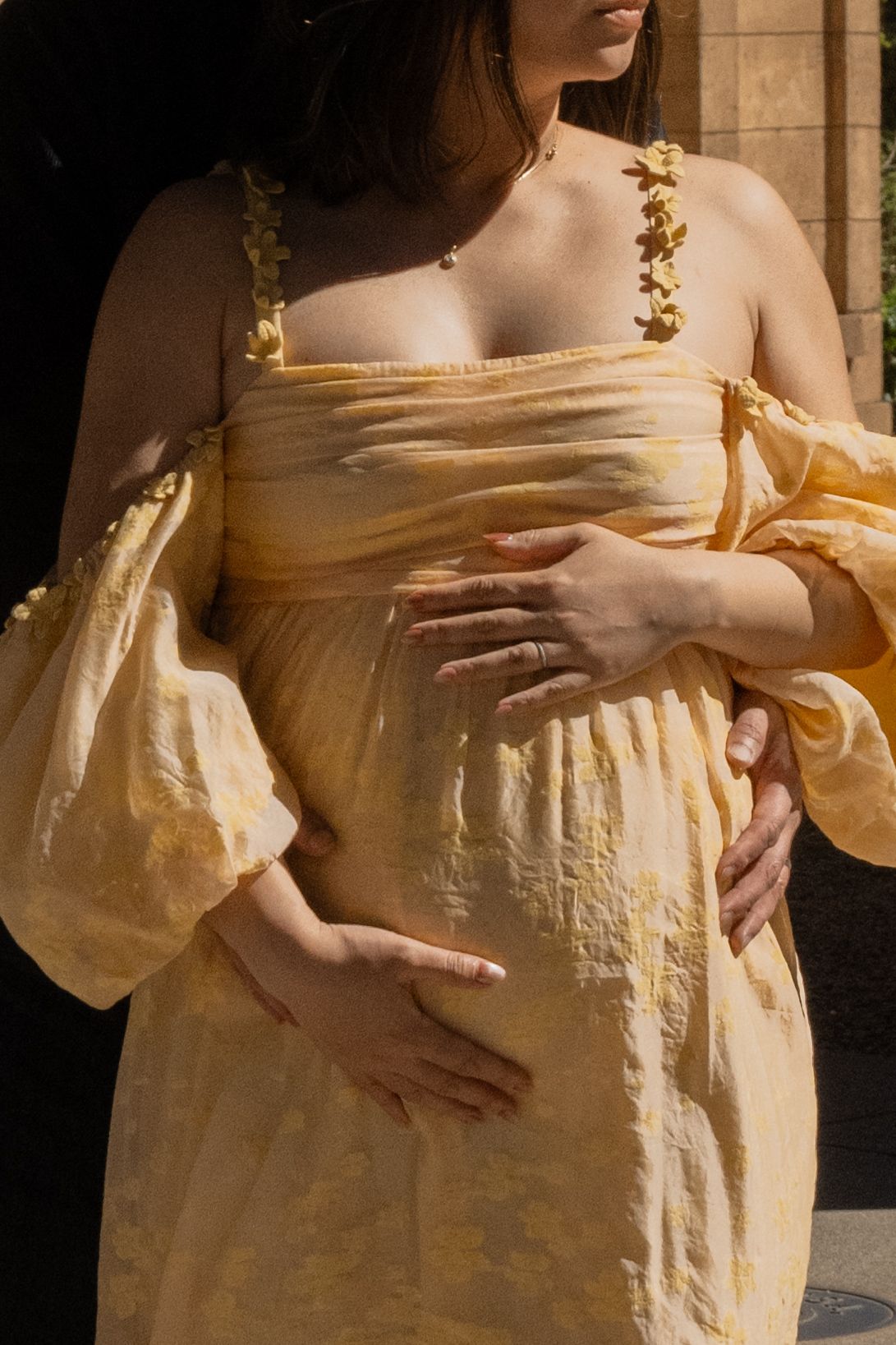 Pregnant woman in yellow flowy dress cradling baby bump in sunlight.