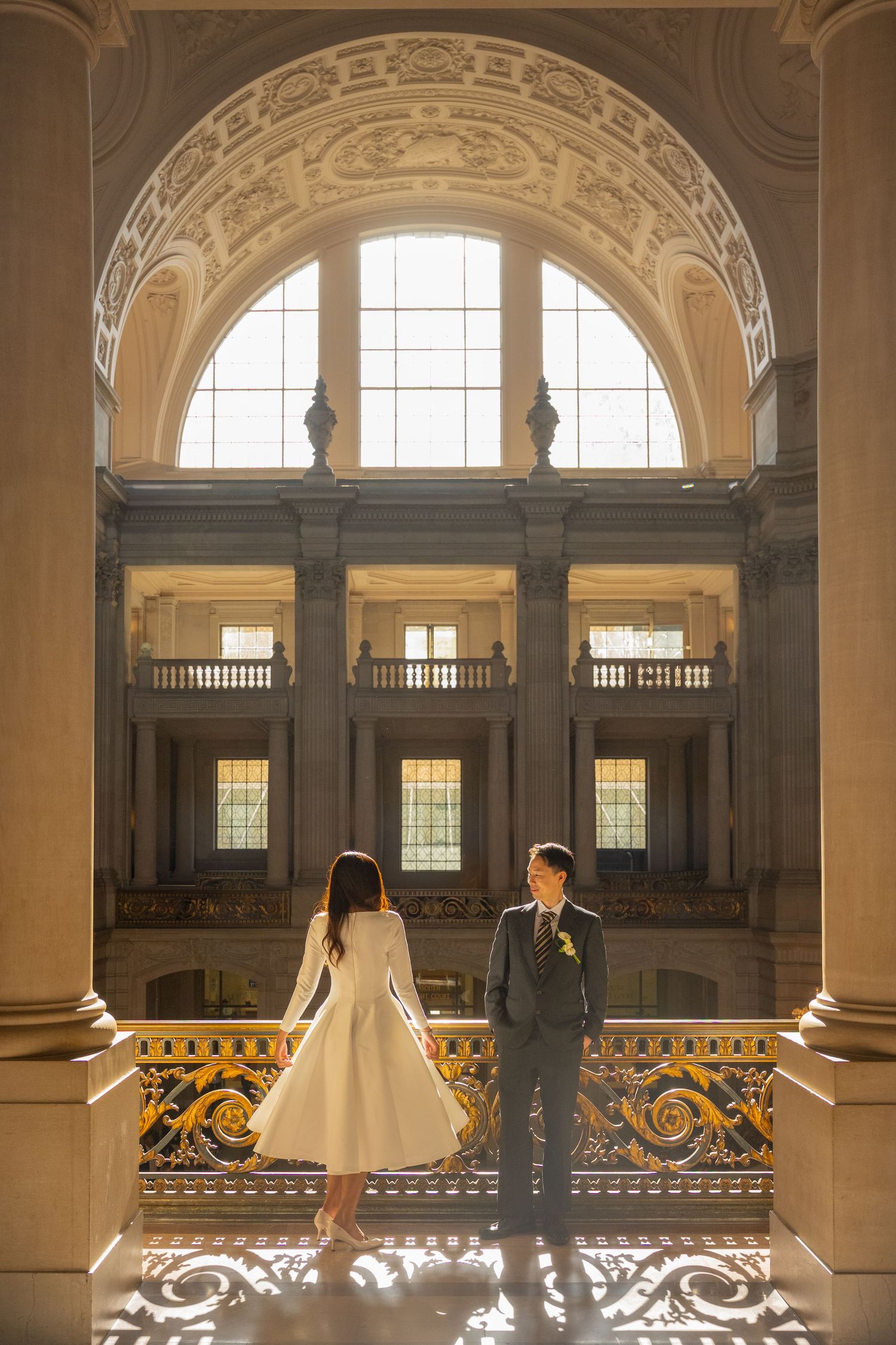 Bride and groom stand on ornate golden balcony inside a grand Beaux-Arts building with arched windows.