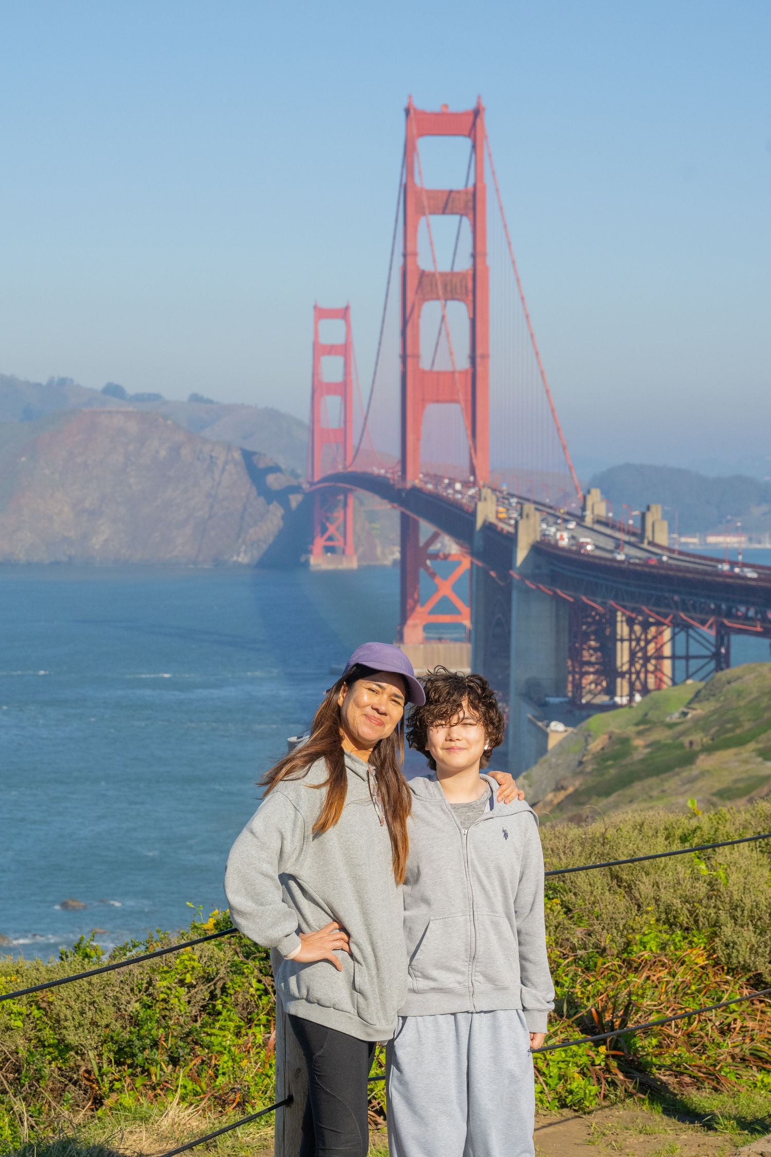 Two teenagers pose together in front of the iconic Golden Gate Bridge on a sunny day.