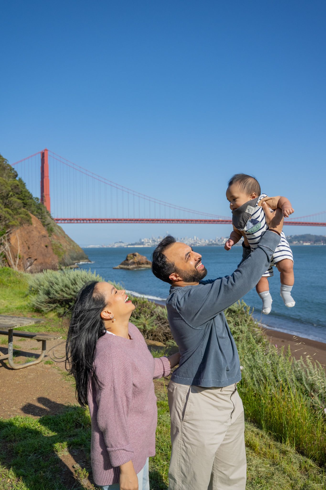 Family lifting baby near Golden Gate Bridge on sunny day.