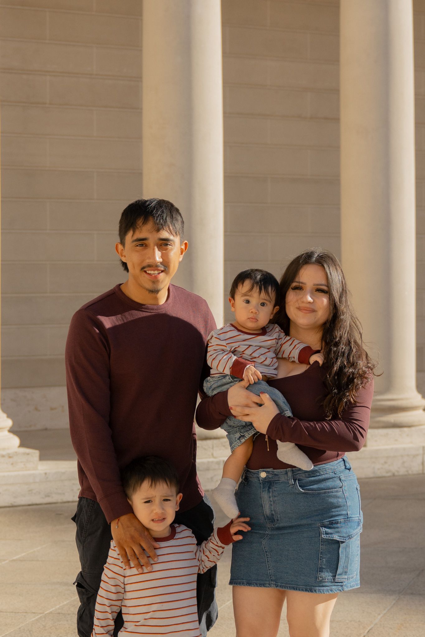 A young family of four poses in front of large stone columns, with parents holding two young children.