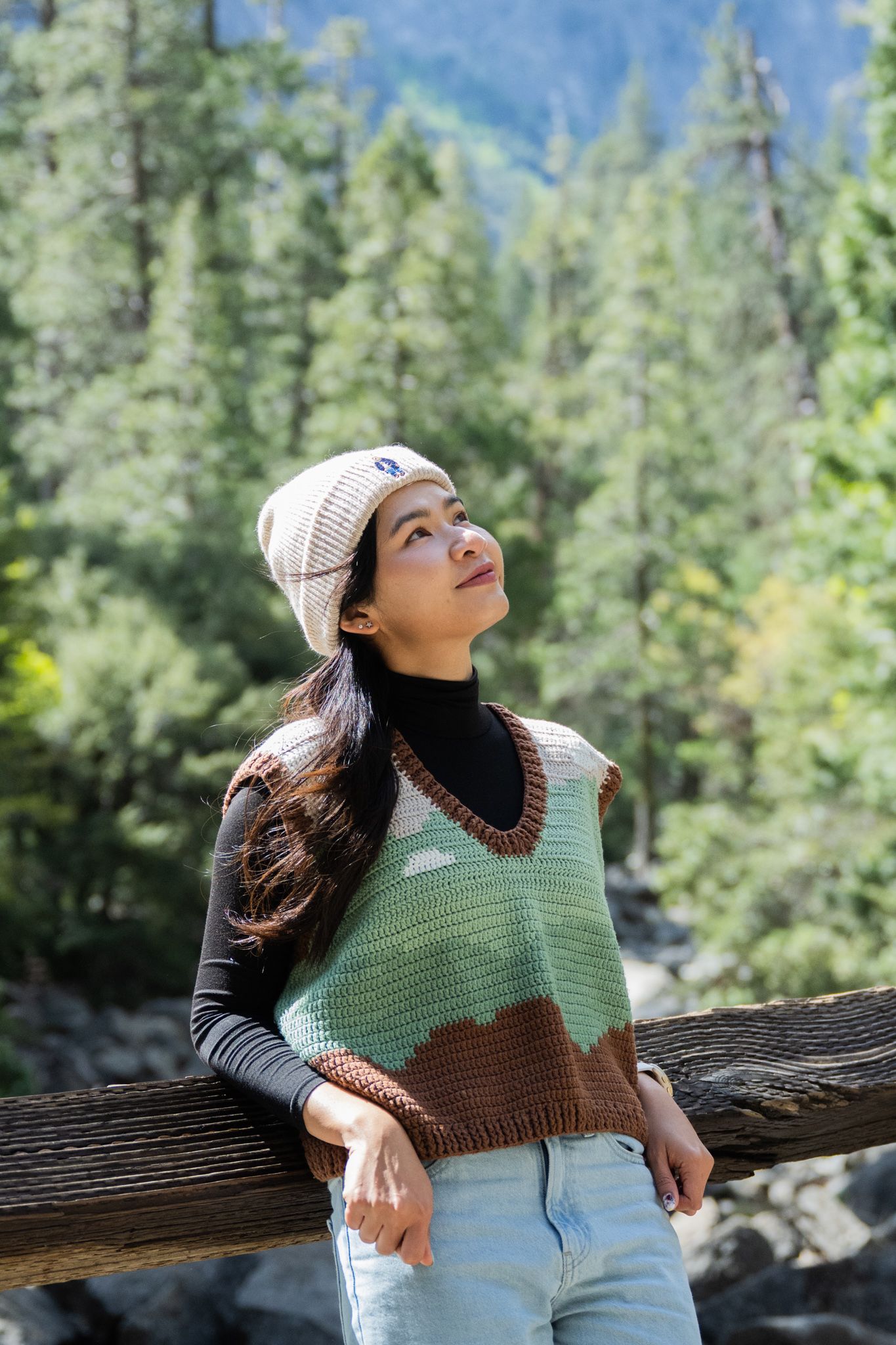 Young woman in knit vest and beanie leans on wooden fence gazing upward in forest setting.