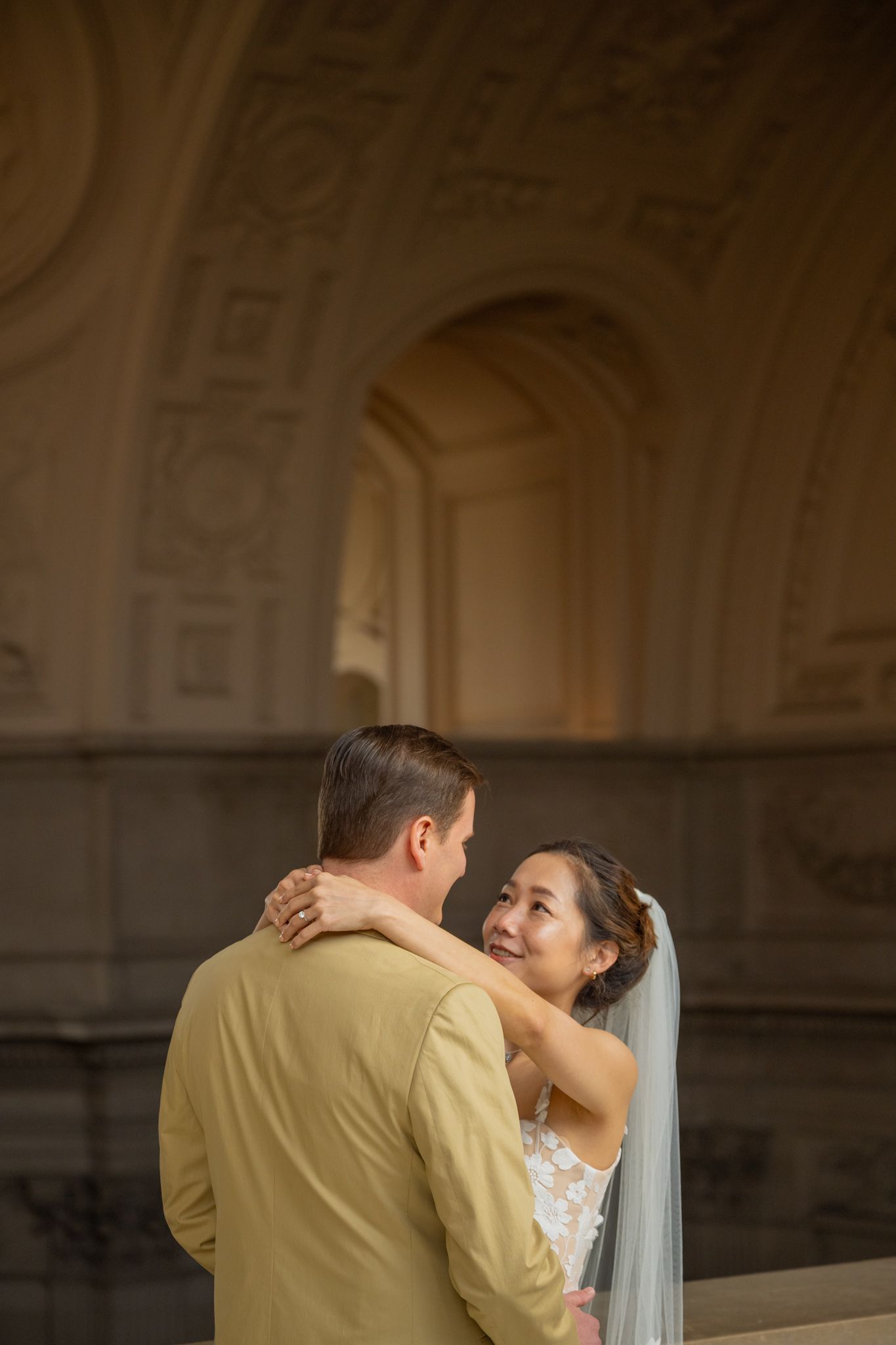 Smiling bride in lace gown embraces groom beneath ornate arched ceiling of elegant venue.