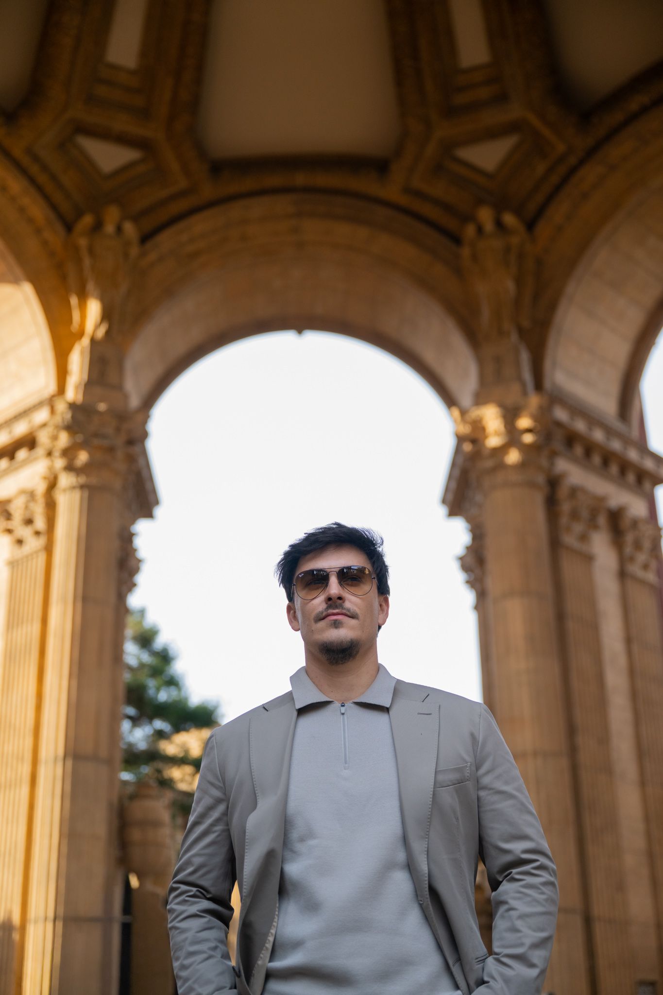 Stylish man in beige blazer and sunglasses stands under ornate stone arches at Palace of Fine Arts.