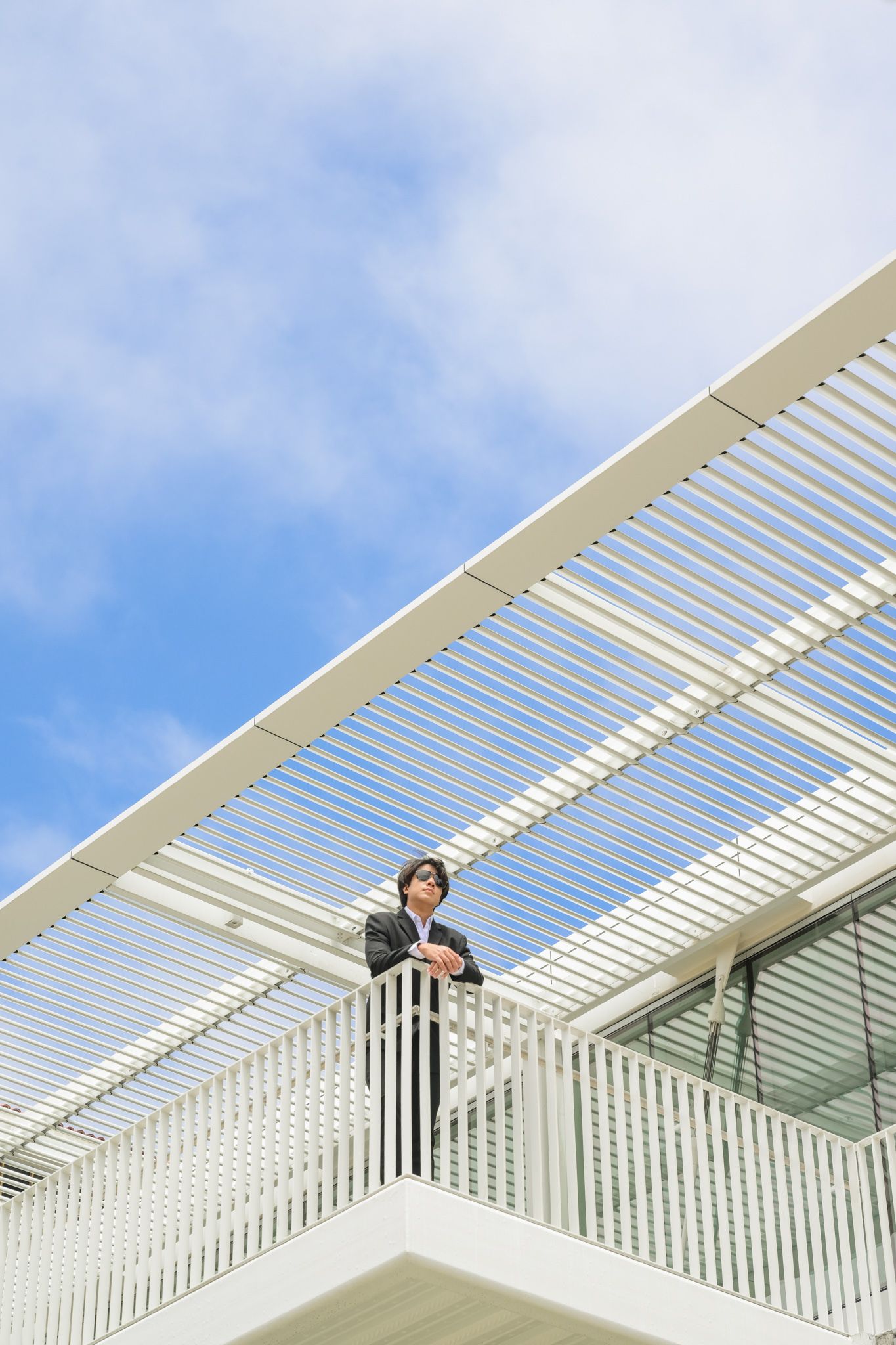 Businessman in suit stands on modern white architectural balcony against a bright blue sky.