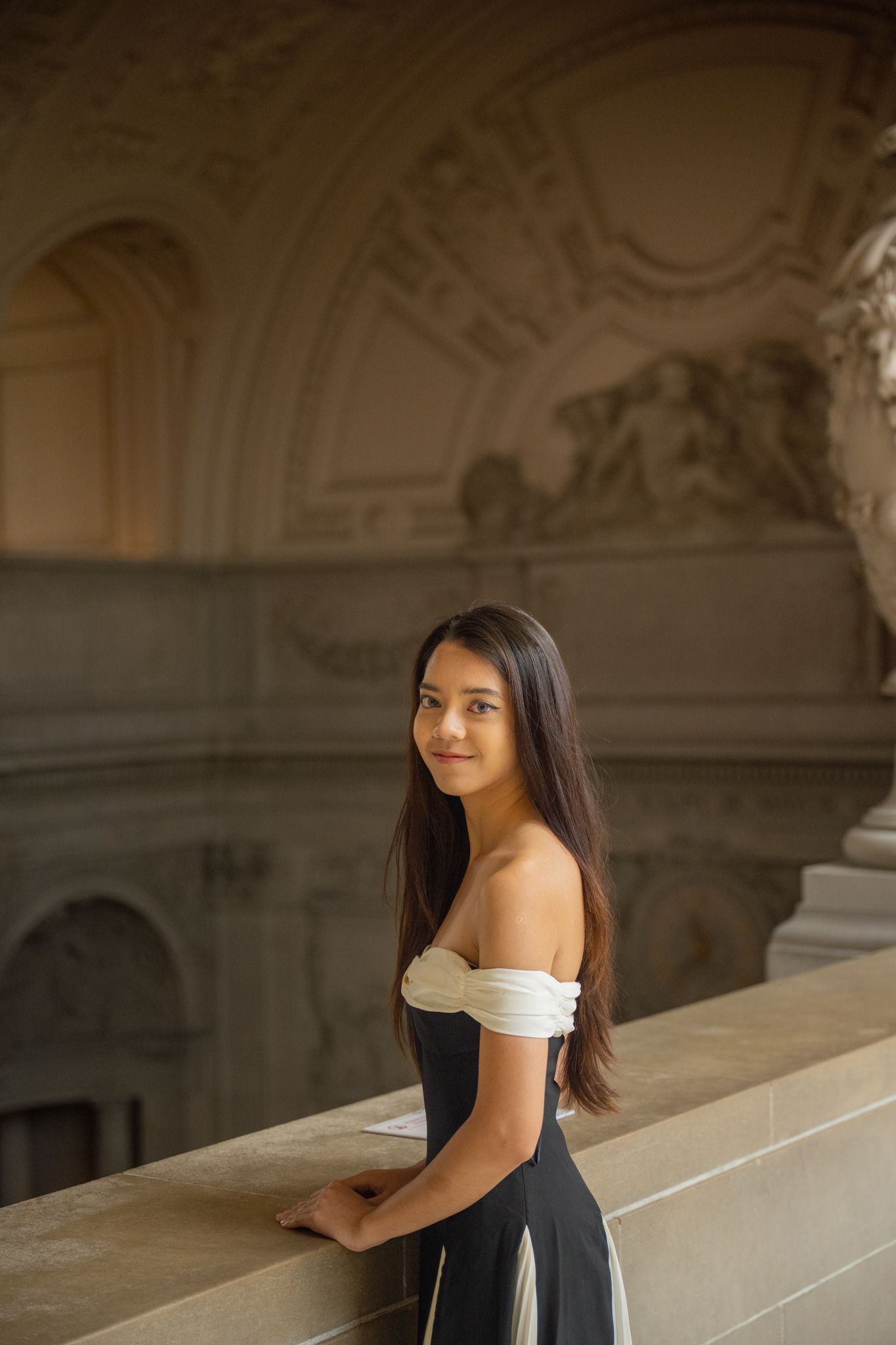 Young woman in elegant off-shoulder black and white dress poses at ornate classical architecture balcony.