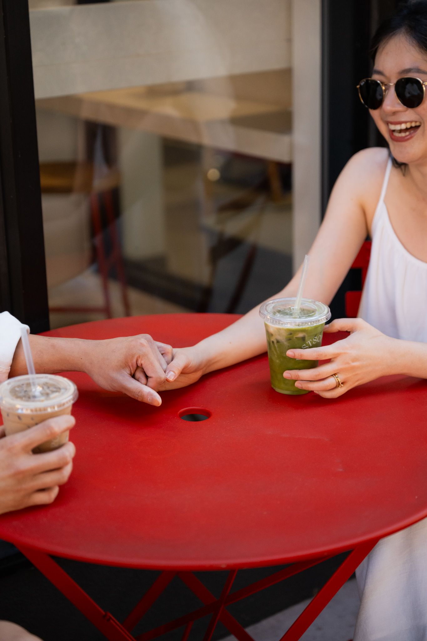 Two women sitting at a red outdoor cafe table, smiling and holding iced green matcha and coffee drinks.