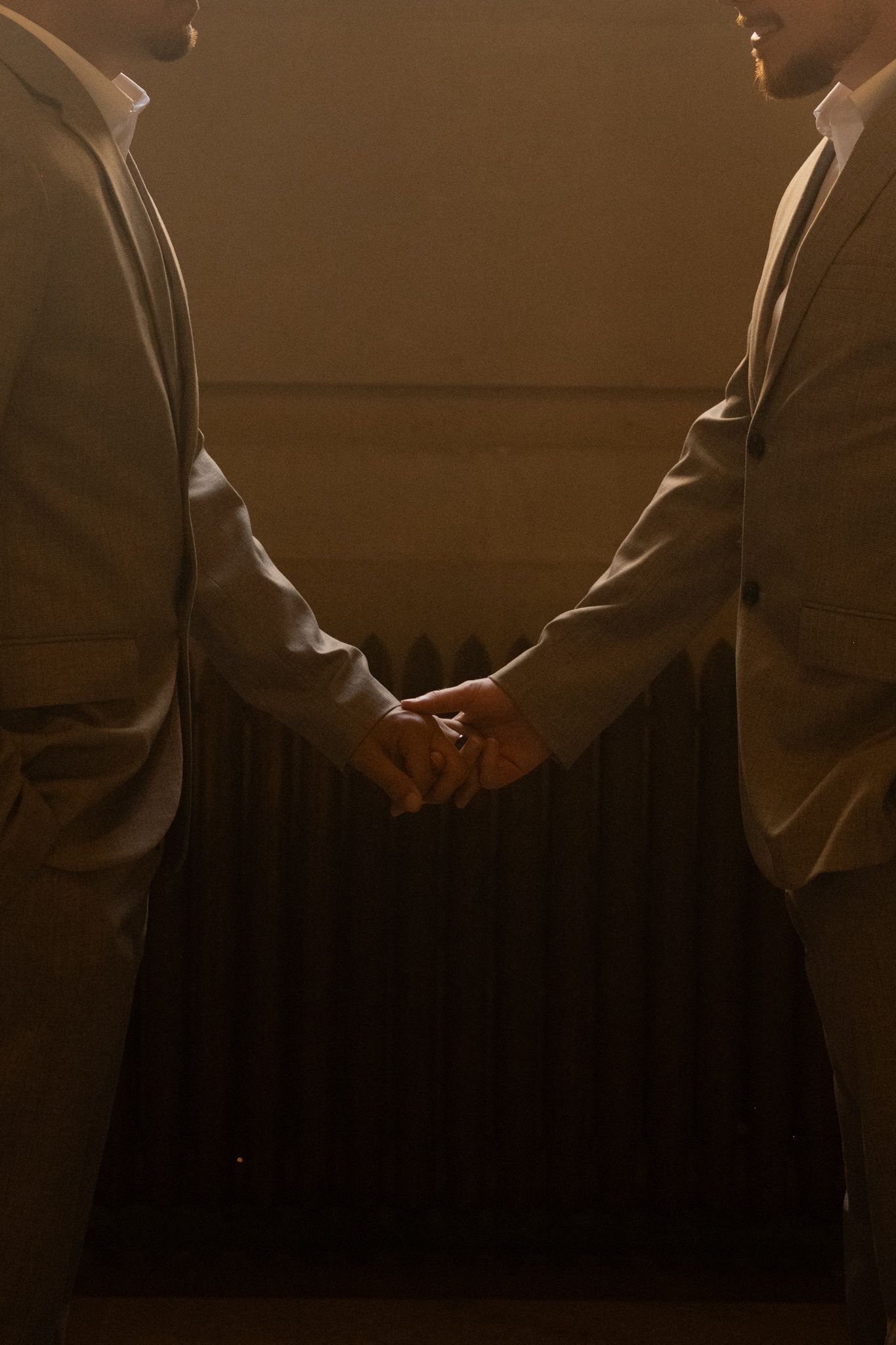 Two grooms in matching gray suits hold hands in a dramatic warmly lit wedding portrait.