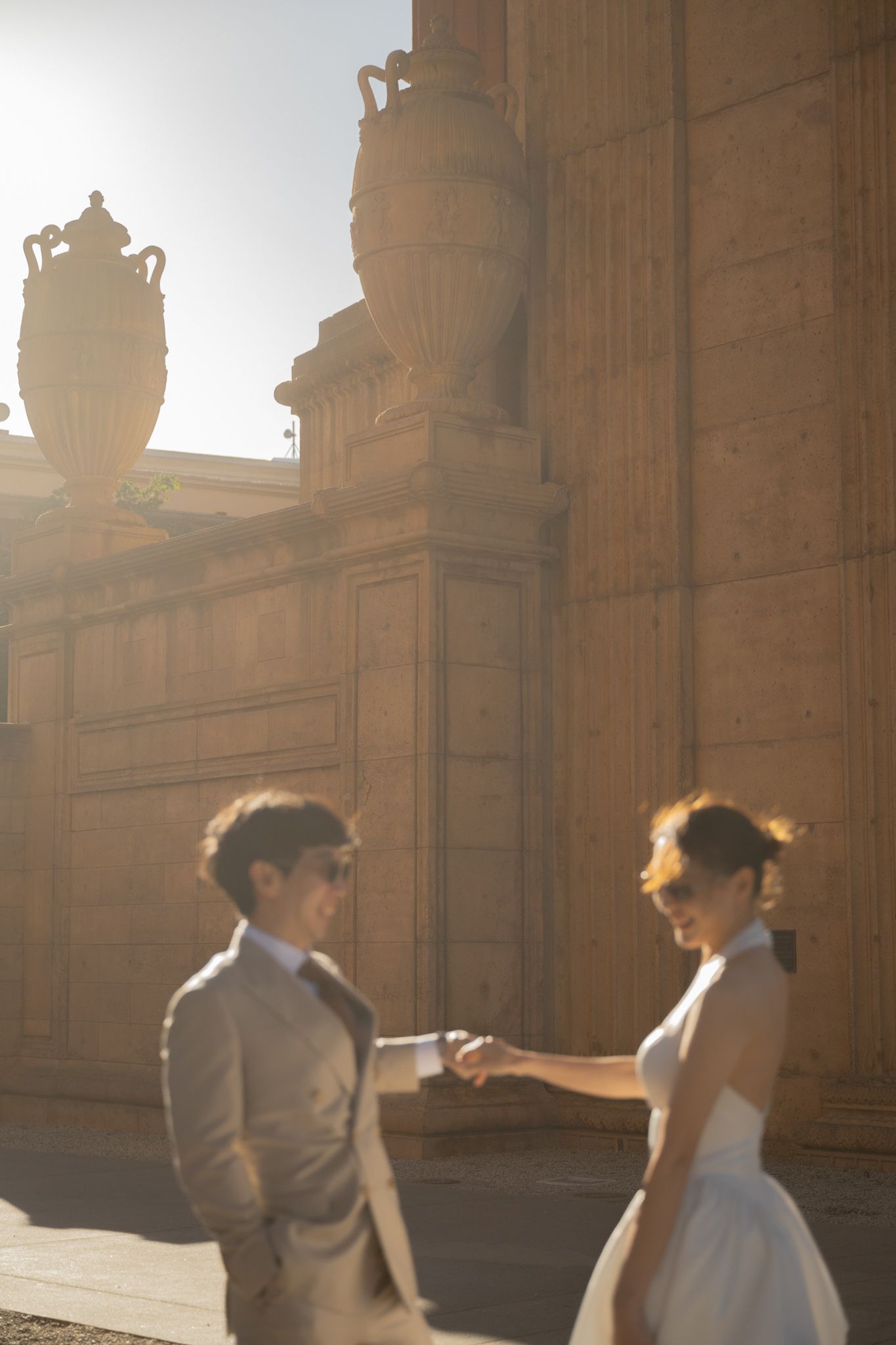 Bride and groom in sunglasses holding hands outside a classical stone building with ornate urns in bright sunlight.