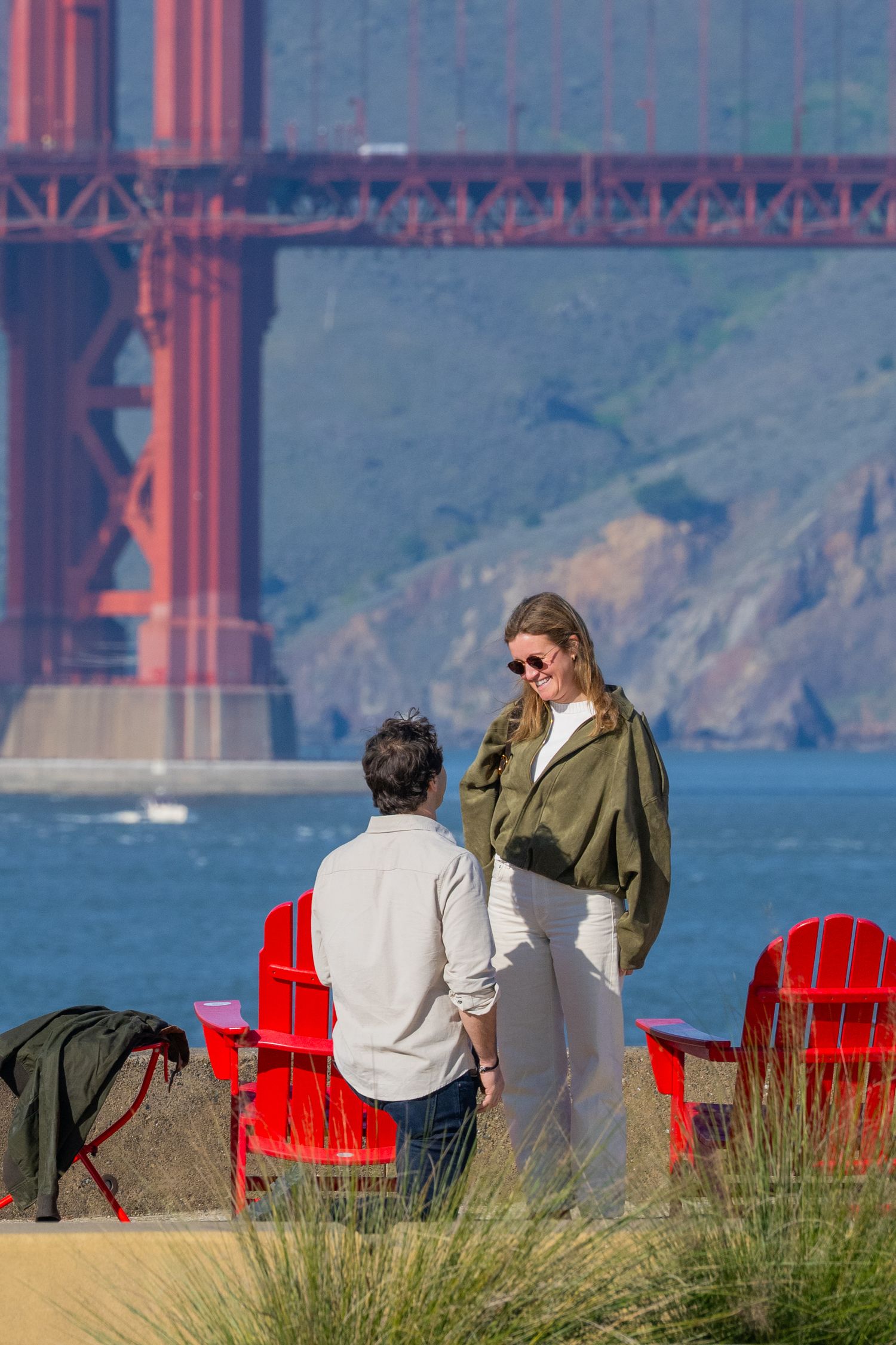 A couple holds hands near red Adirondack chairs with the Golden Gate Bridge in the background.
