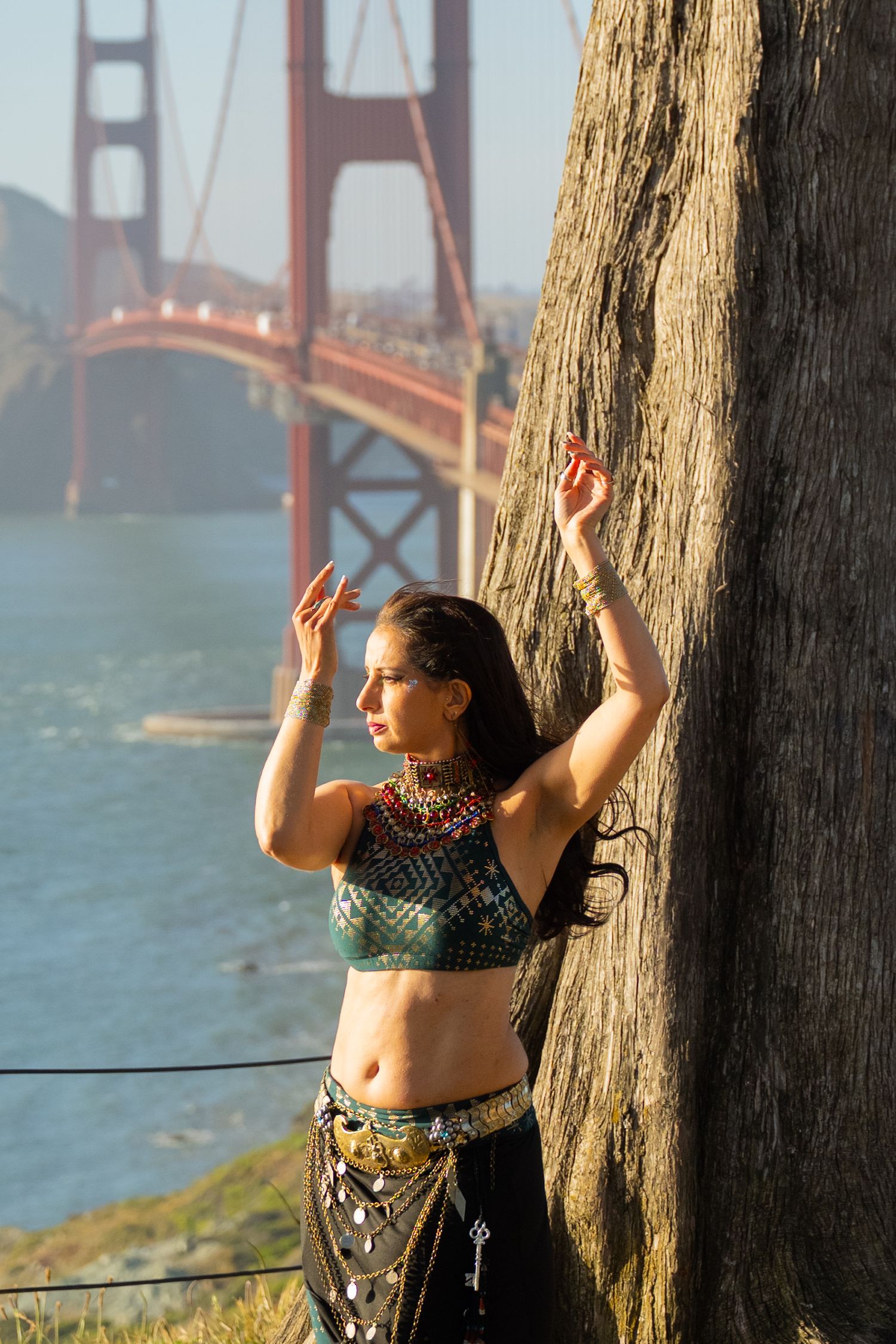 A woman in tribal belly dance costume poses gracefully by a tree with the Golden Gate Bridge behind her.