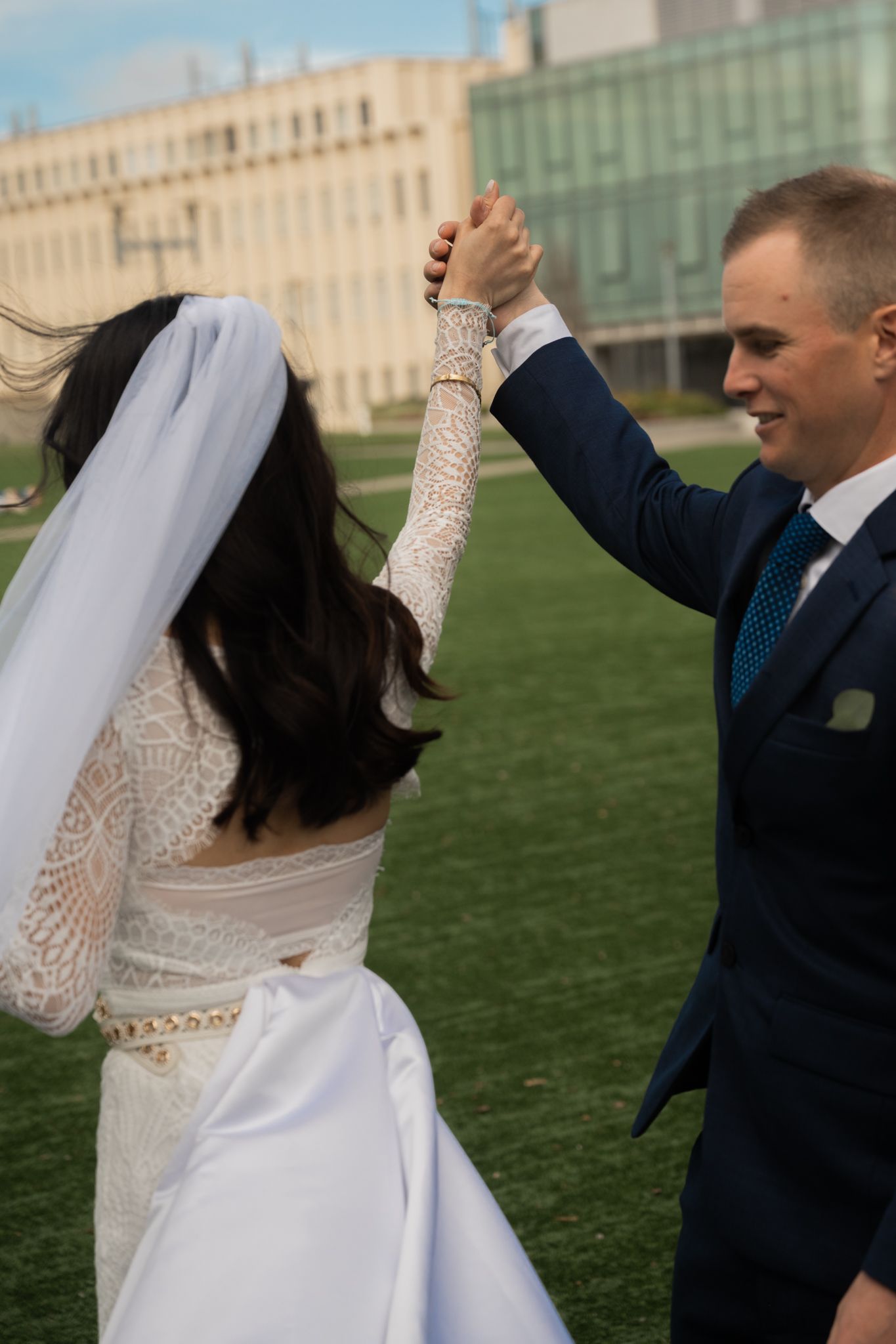 Groom in navy suit twirls laughing bride in lace wedding dress on green lawn outdoors.