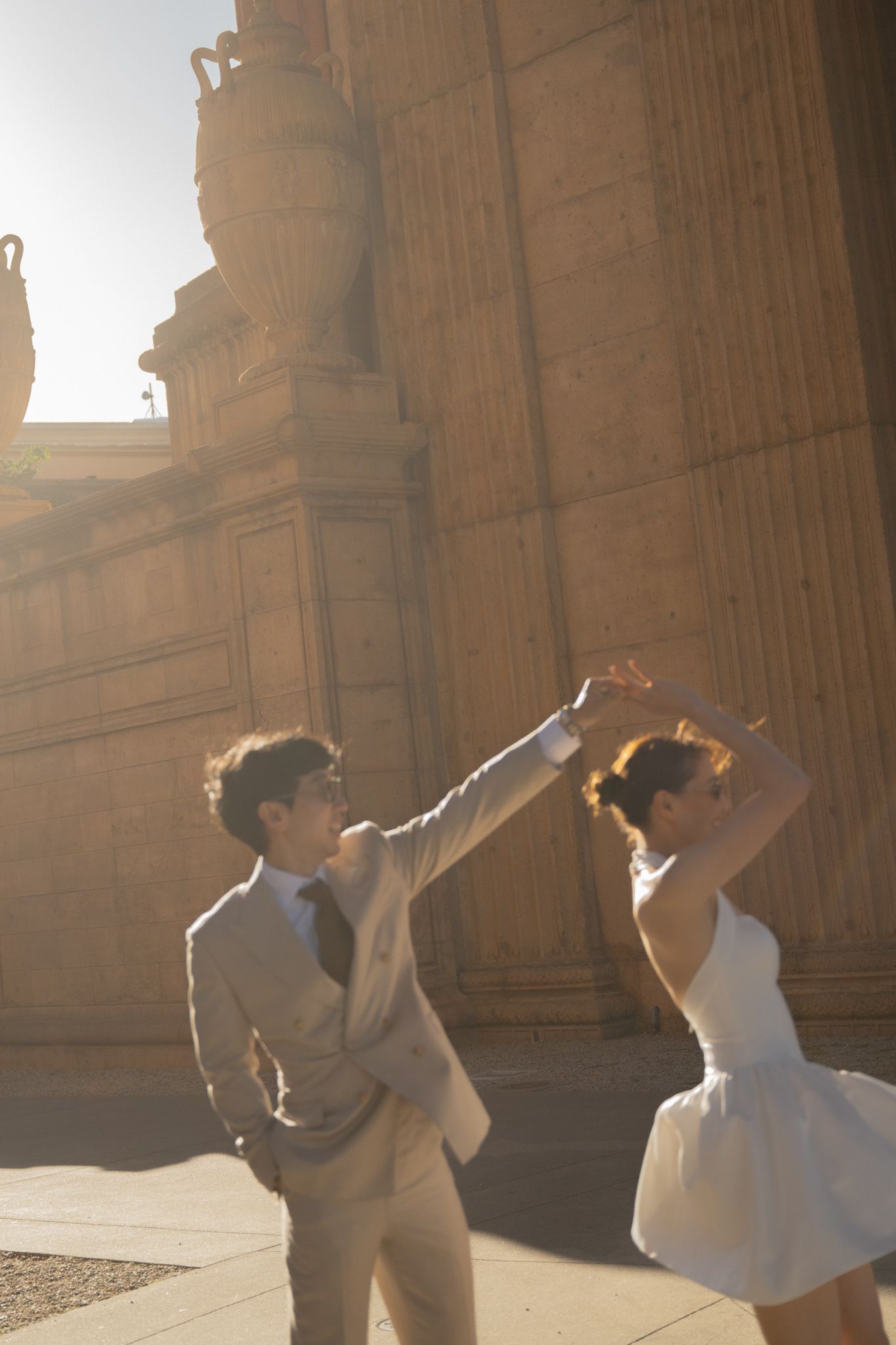 Bride and groom spin and dance joyfully in warm sunlight beside grand stone columns of a historic building.