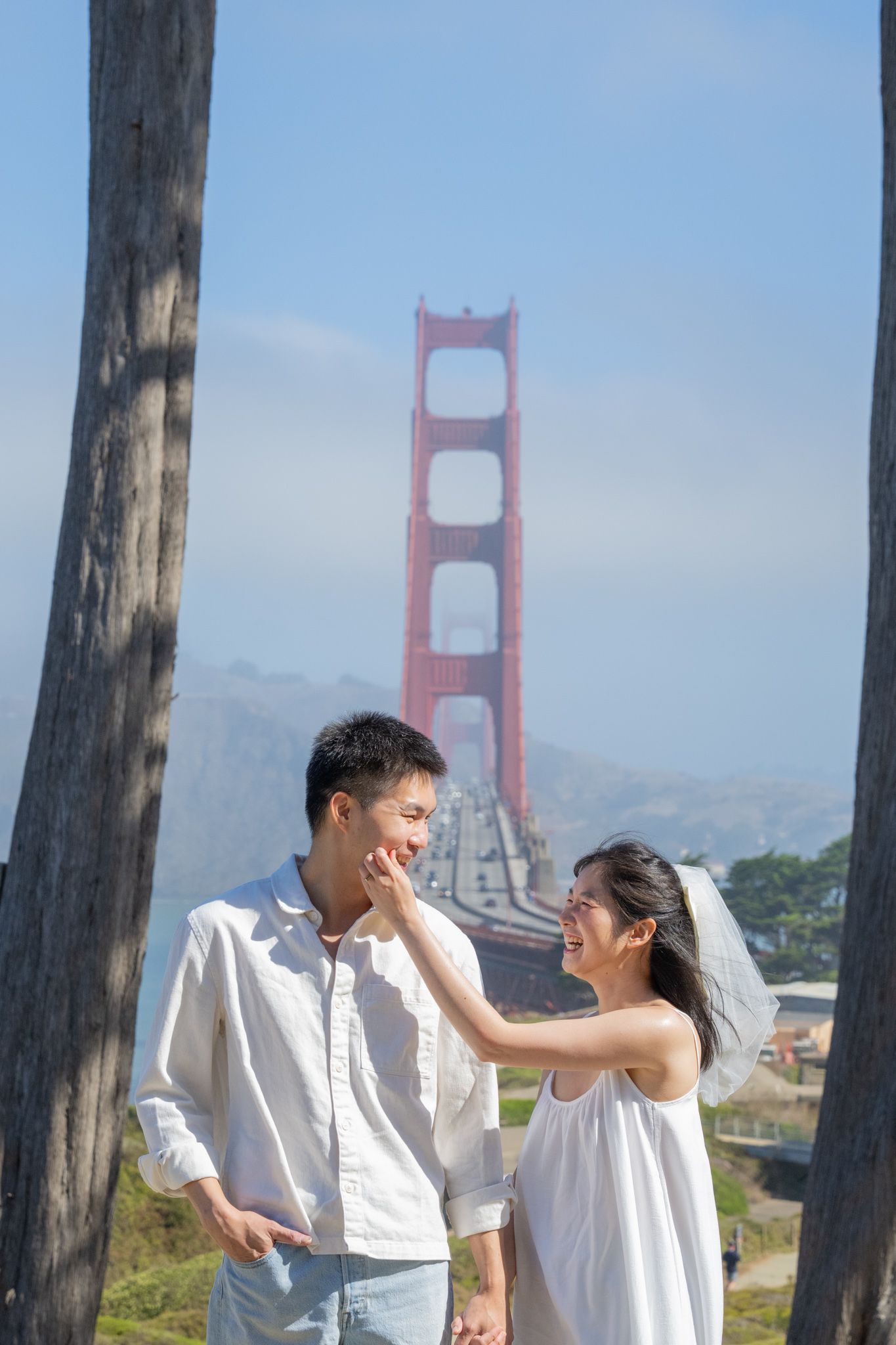 Couple in white outfits smiling and holding hands near Golden Gate Bridge on a sunny day.
