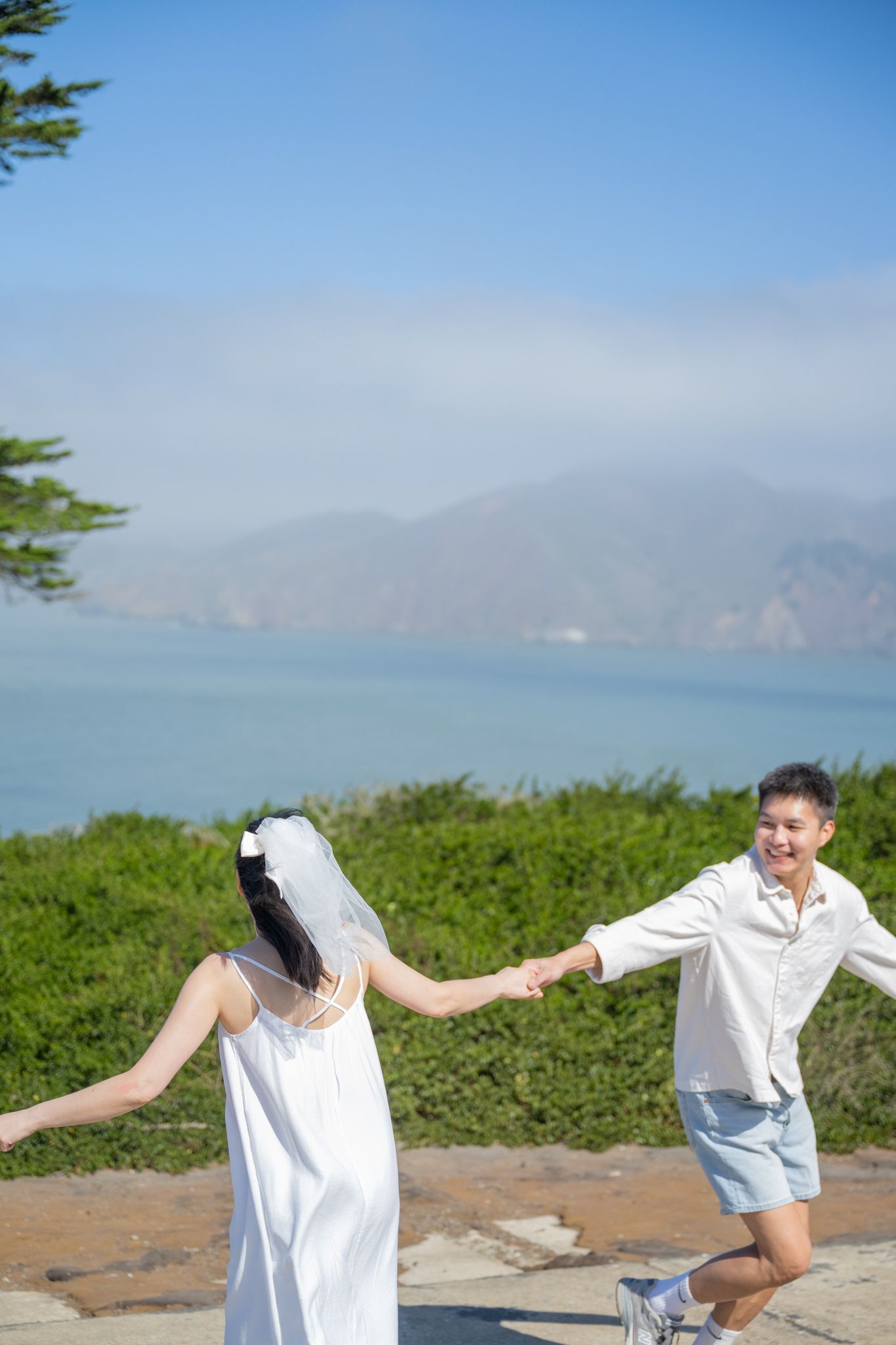 A joyful couple in wedding attire holds hands outdoors, with scenic ocean and mountain views under a clear blue sky.