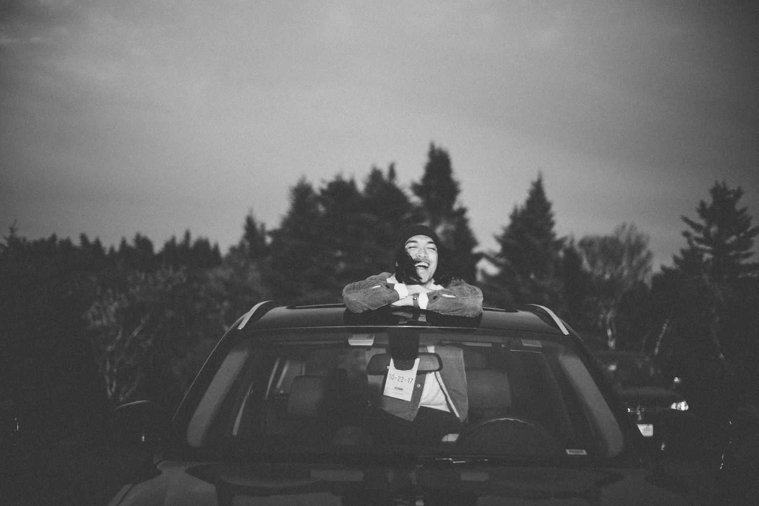 Black and white photo of person emerging from car sunroof surrounded by tall evergreen trees at dusk.