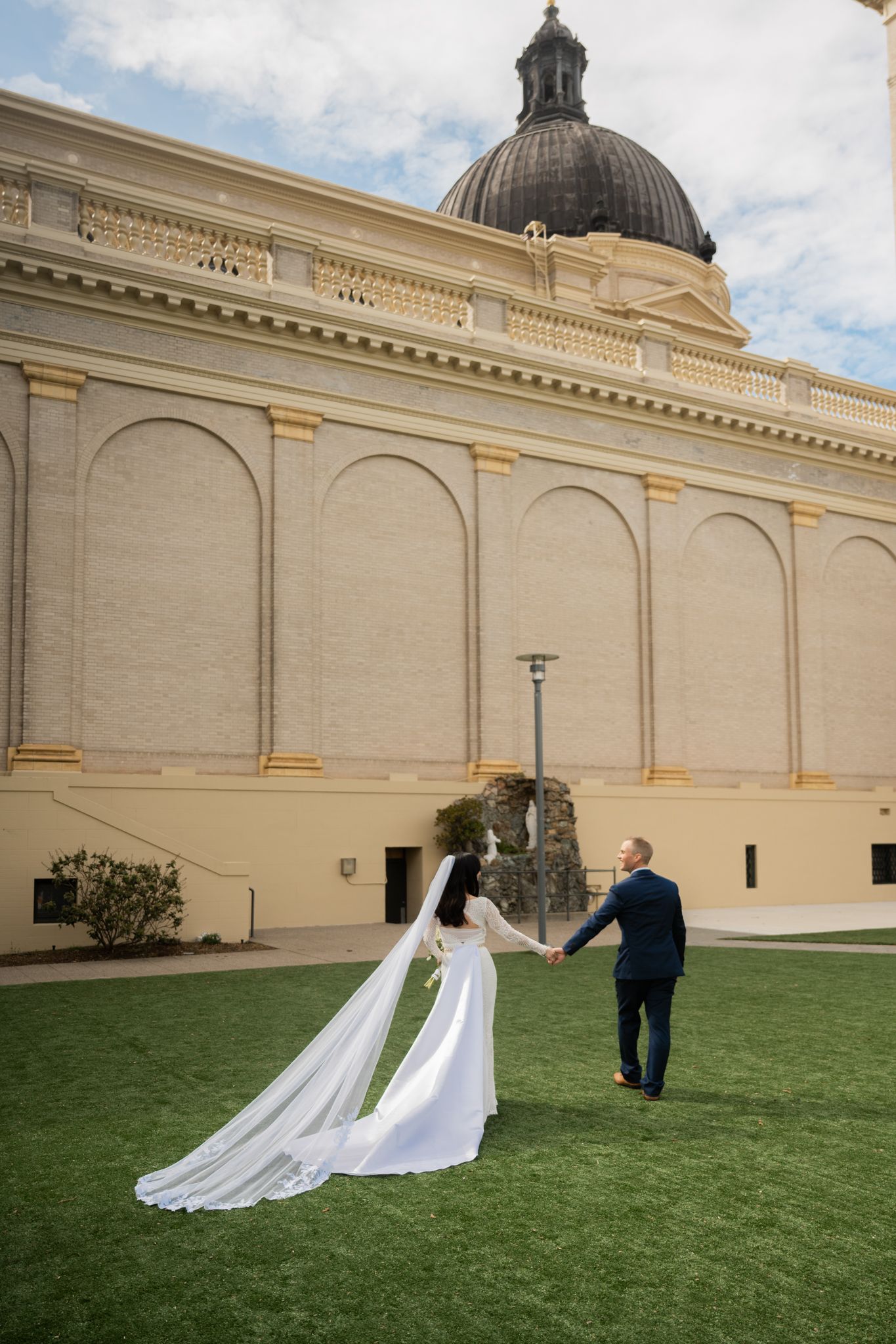 Bride and groom hold hands walking on green lawn before grand domed neoclassical building exterior.